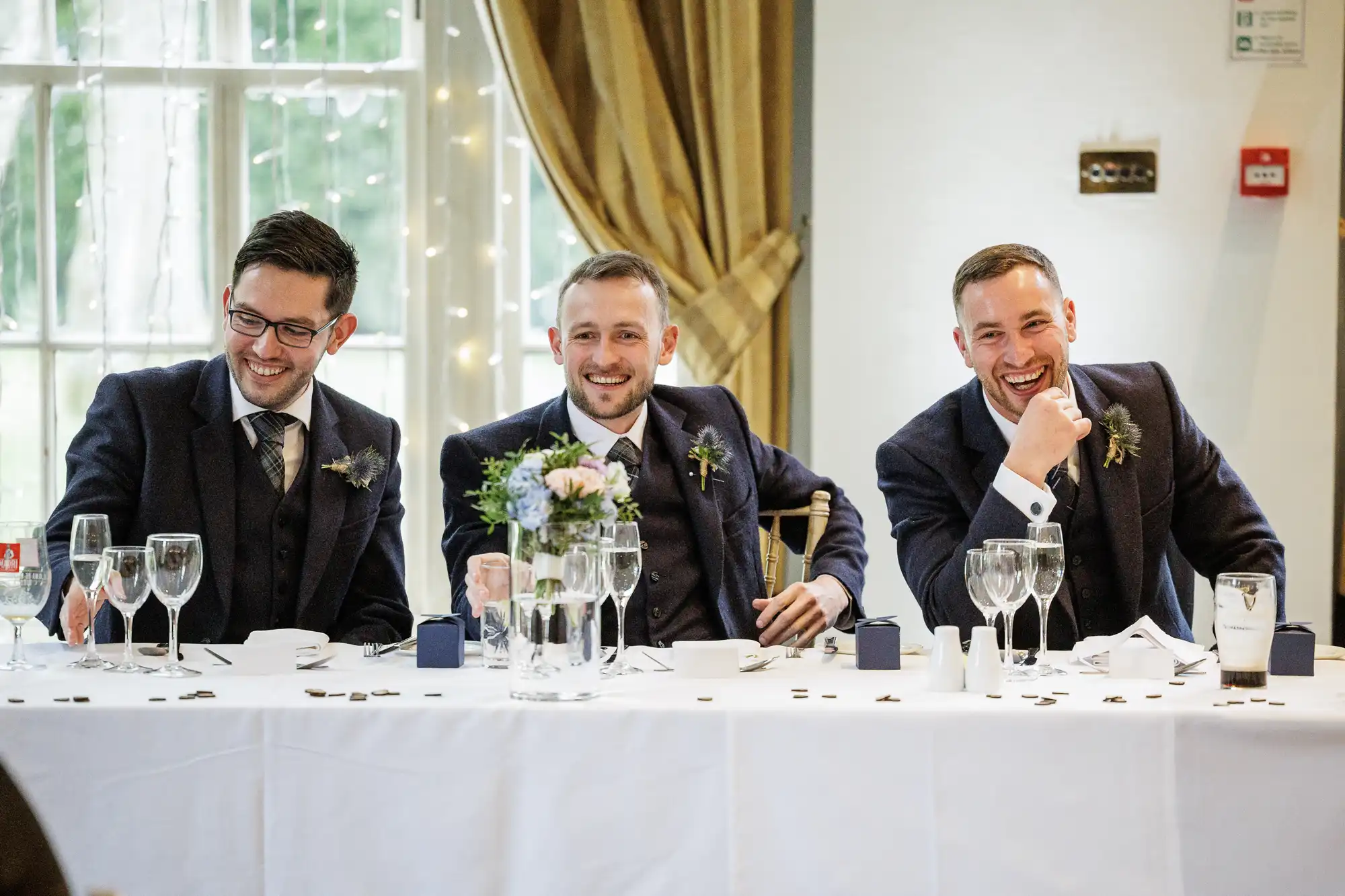 Three men in dark suits sit at a long white table set with glasses and flowers, smiling and laughing. They are indoors, in front of a large window with curtains and fairy lights, suggesting a festive or formal event such as a wedding reception.