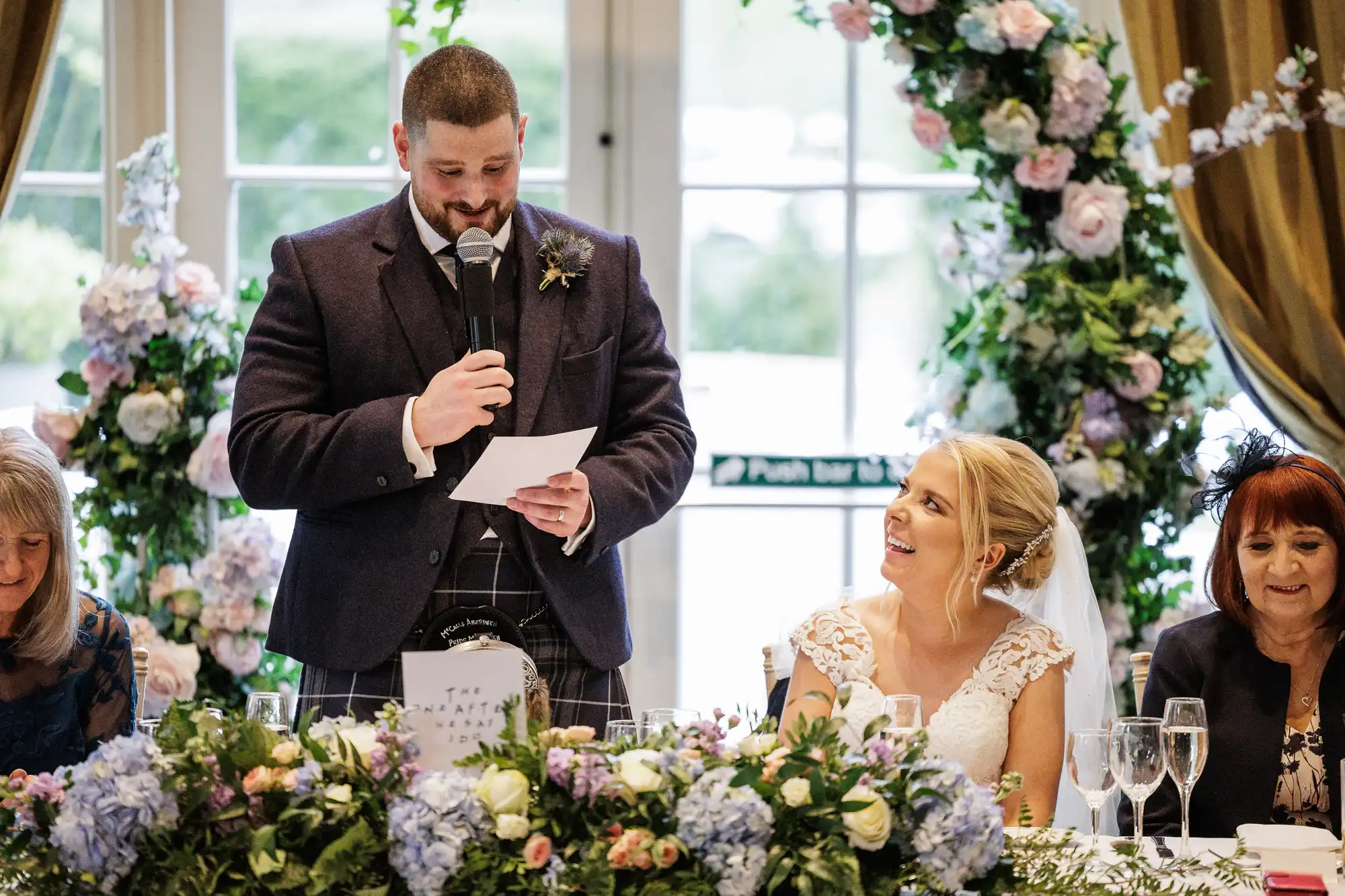 A man in a suit stands with a microphone and reading notes, giving a speech at a wedding reception. A bride in a white dress smiles at him. Floral arrangements and greenery decorate the table and background. Two women sit on either side.