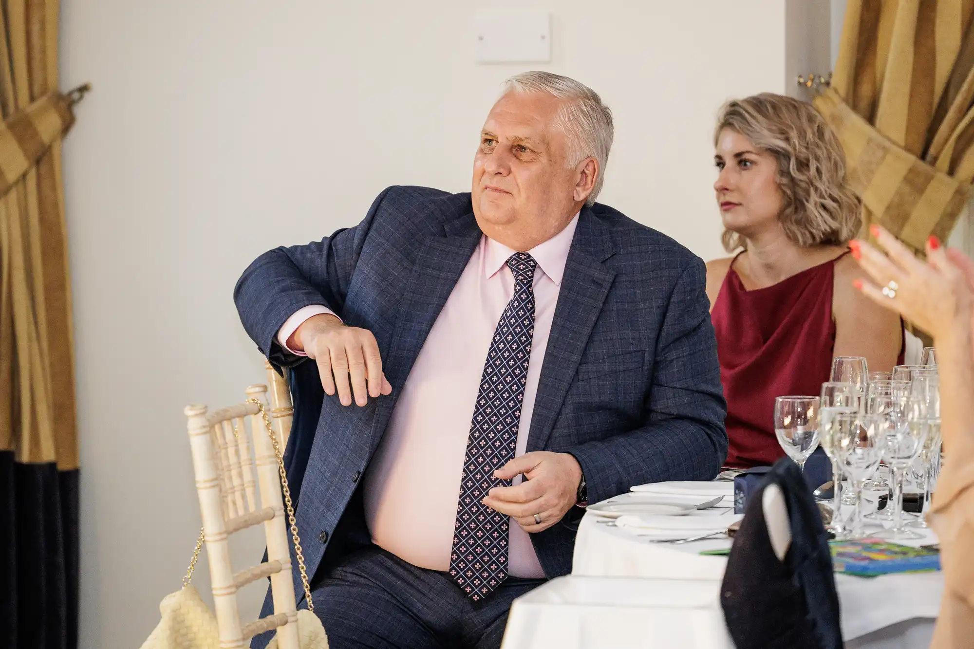 An older man in a blue suit and pink shirt sits at a table set for a formal event, with a woman in a red dress beside him. Both look towards the right, appearing attentive. Gold curtains hang in the background and glassware is arranged on the table.