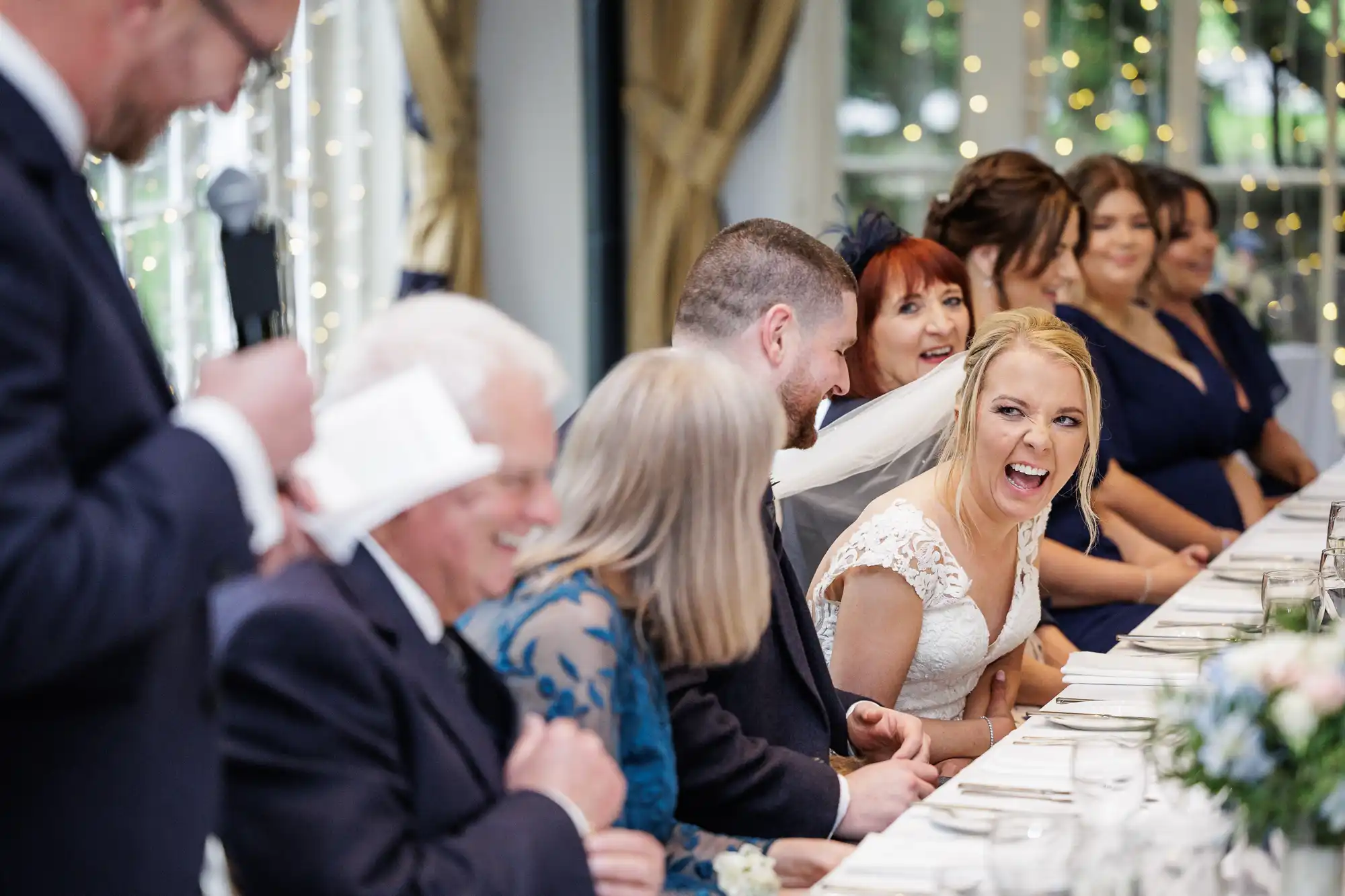 A joyful bride in a white dress laughs at a long table during a wedding reception, surrounded by smiling guests in formal attire. Fairy lights and draped curtains decorate the background, creating a festive and warm atmosphere.