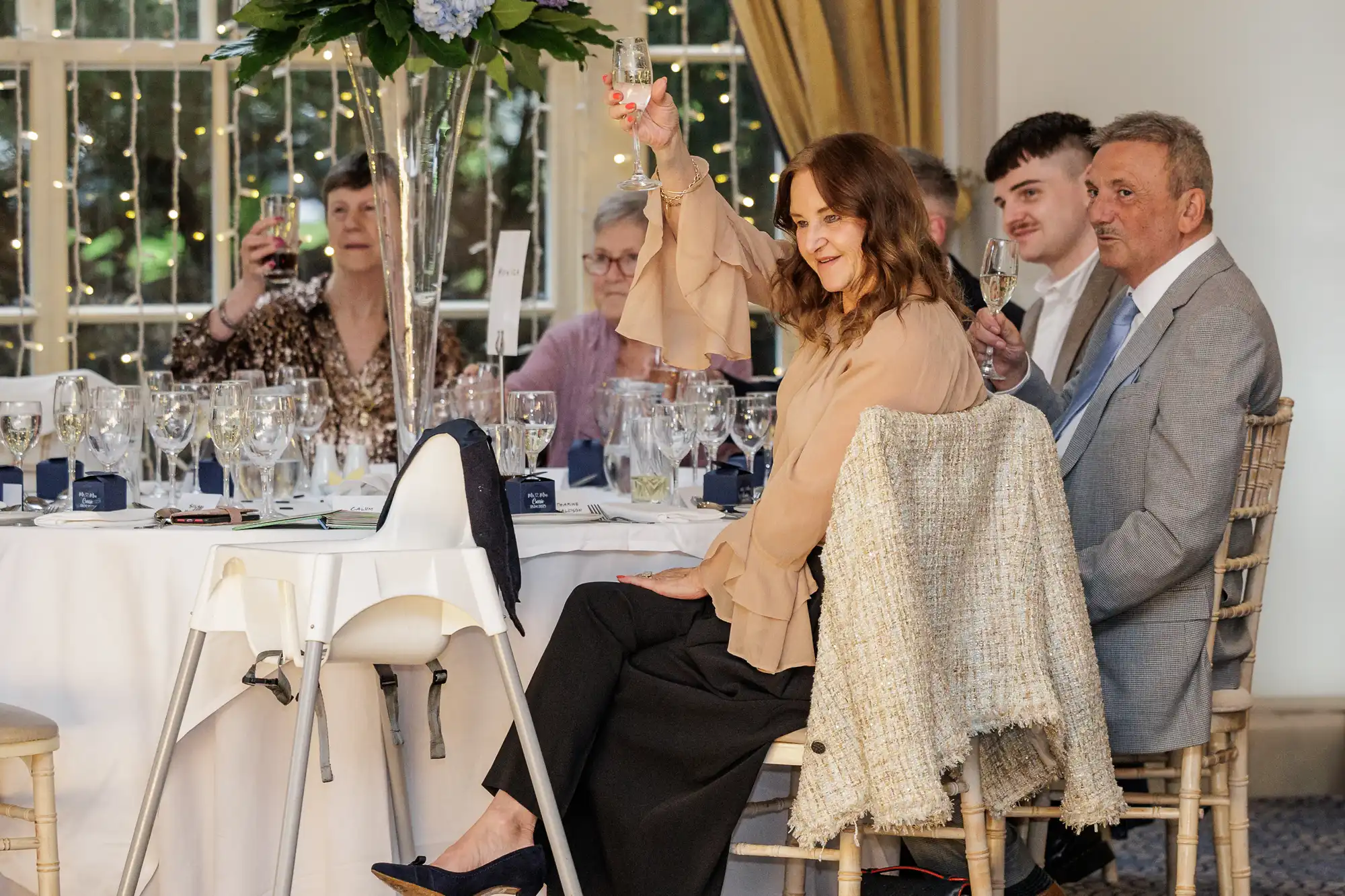A group of people sit around a round banquet table, raising glasses in a toast. The table is elegantly set with wine glasses and flowers. Warm lights and a window are in the background. A woman in a beige blouse smiles and faces the camera, mid-toast.