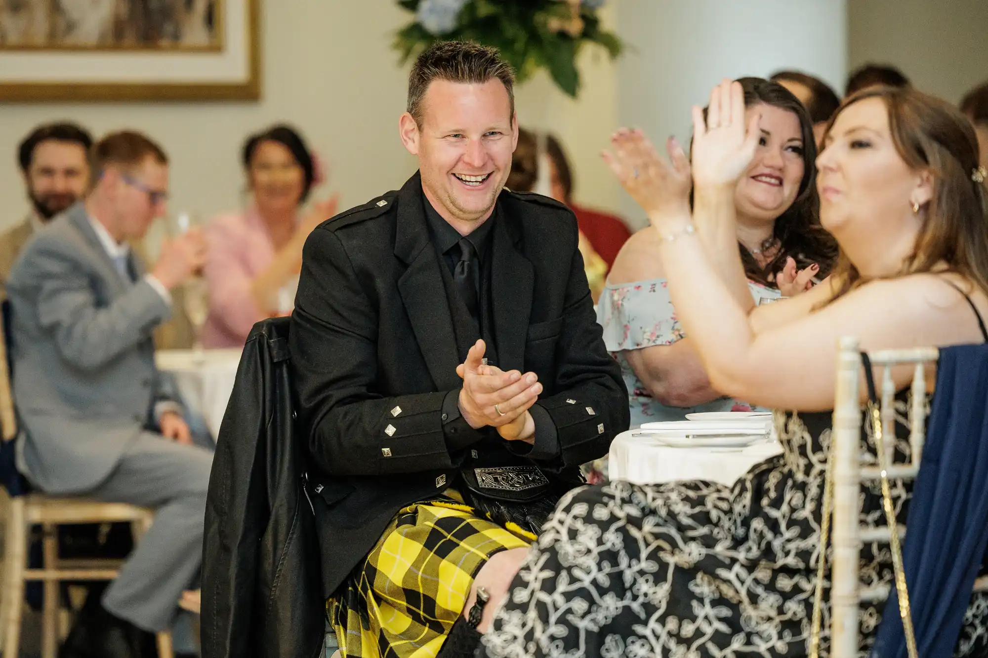 A man in a black jacket and yellow tartan kilt smiles and claps at an event. He sits at a table among other seated guests, some of whom are also applauding. The background shows more people, a painting, and floral decorations.