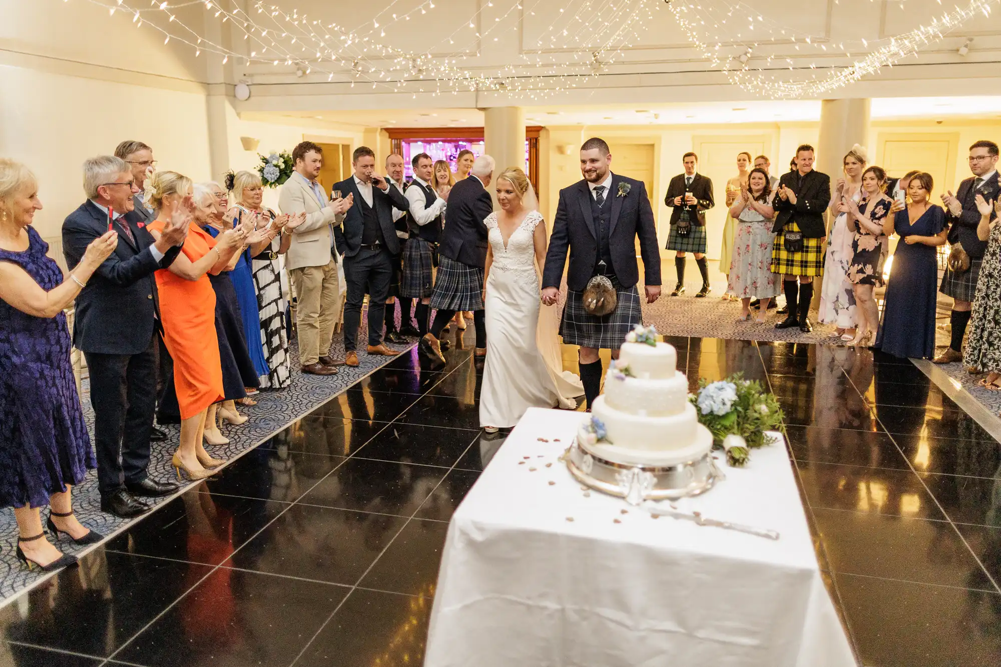 A bride and groom hold hands, smiling as they approach a white wedding cake on a table. Guests in formal attire stand around them, clapping and cheering. The room is decorated with fairy lights overhead, creating a festive and joyful atmosphere.
