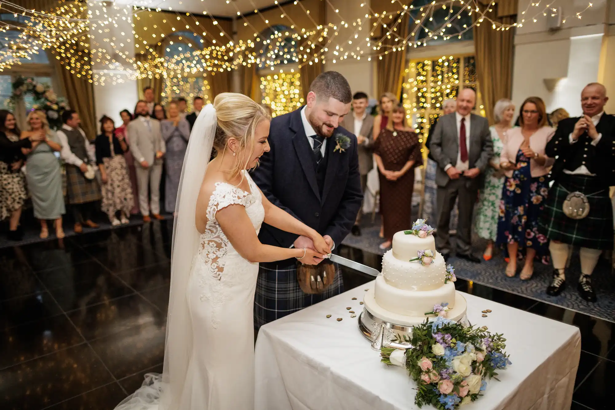 A bride in a white dress and veil and a groom in a dark suit cut a white, two-tier wedding cake together. They smile as guests watch. The room is decorated with gold fairy lights and flowers, creating a festive, joyful atmosphere.