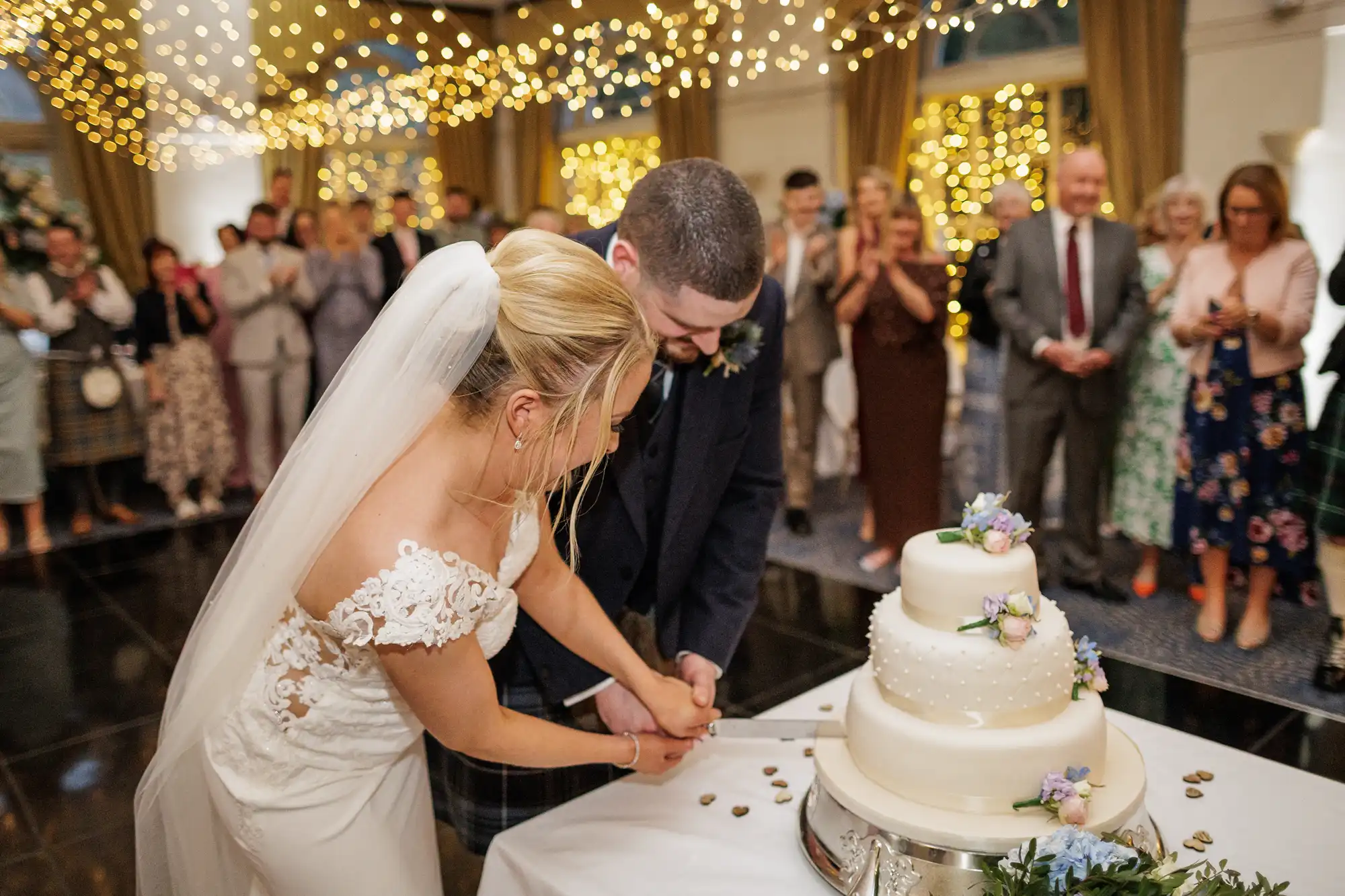 A bride in a white dress and veil and a groom in a dark suit cut a white, three-tiered wedding cake decorated with flowers. Guests stand around them, clapping and smiling, under a ceiling covered with glowing fairy lights.