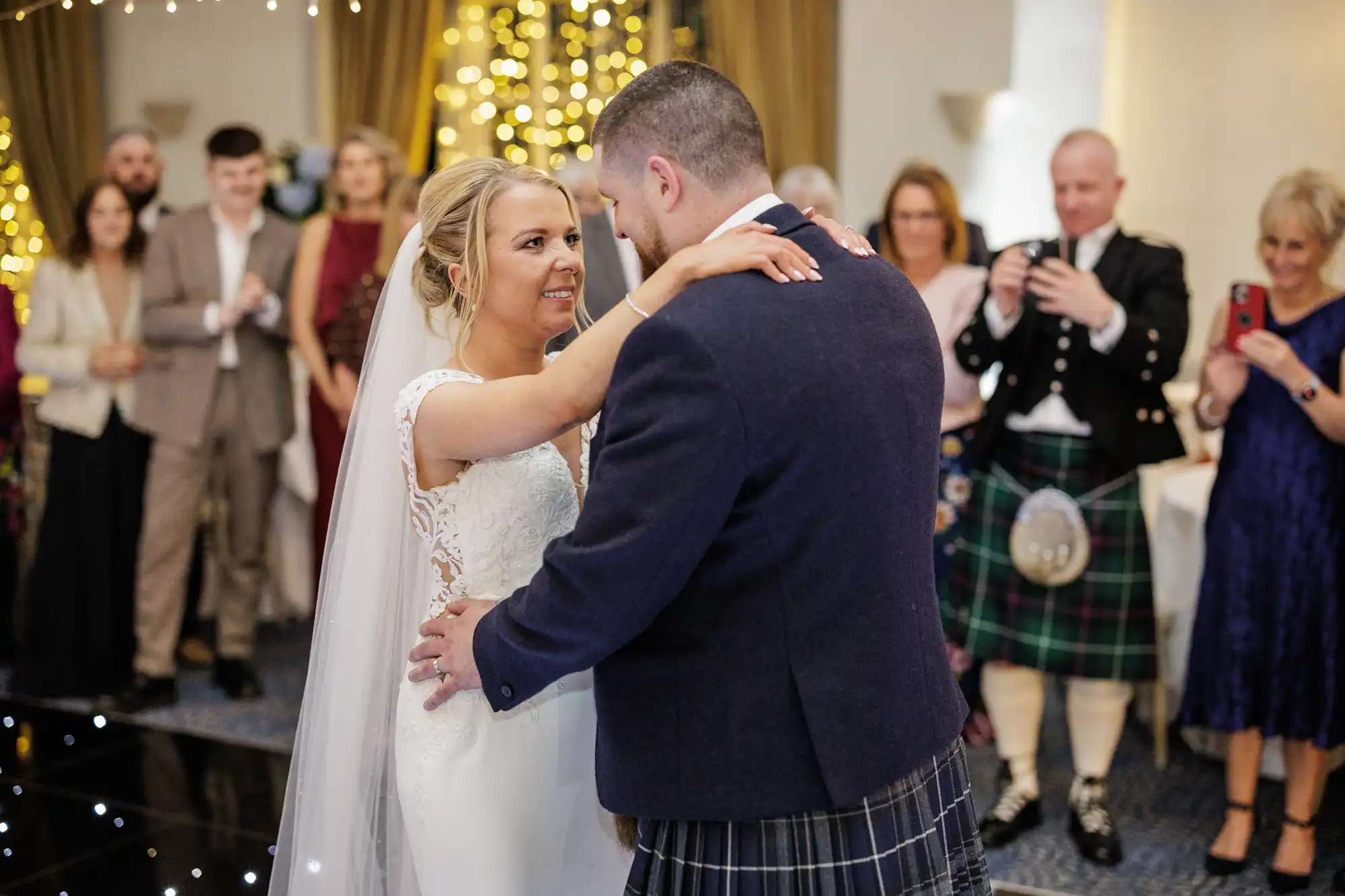 A bride and groom share their first dance at a wedding. The bride wears a white dress and veil, the groom wears a dark jacket and tartan kilt. Guests in formal attire watch, some taking photos. Warm fairy lights and gold drapes decorate the background.