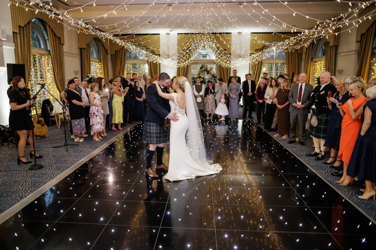 A bride and groom share their first dance on a black, star-lit floor at a wedding reception. Guests stand in a wide circle, watching and smiling. The room is decorated with fairy lights, and some guests are taking photos. The groom wears a kilt, the bride a white dress.