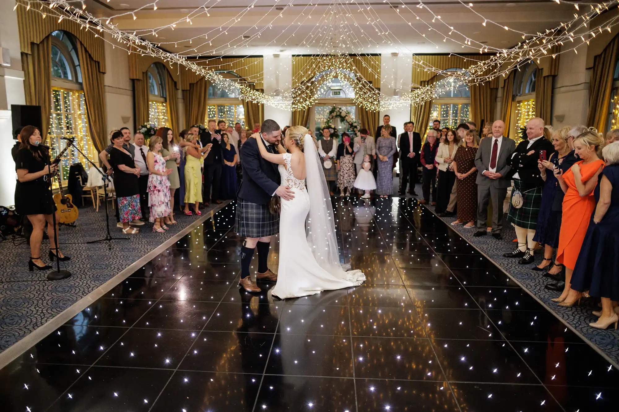 A bride and groom share their first dance on a black, star-lit floor at a wedding reception. Guests stand in a wide circle, watching and smiling. The room is decorated with fairy lights, and some guests are taking photos. The groom wears a kilt, the bride a white dress.
