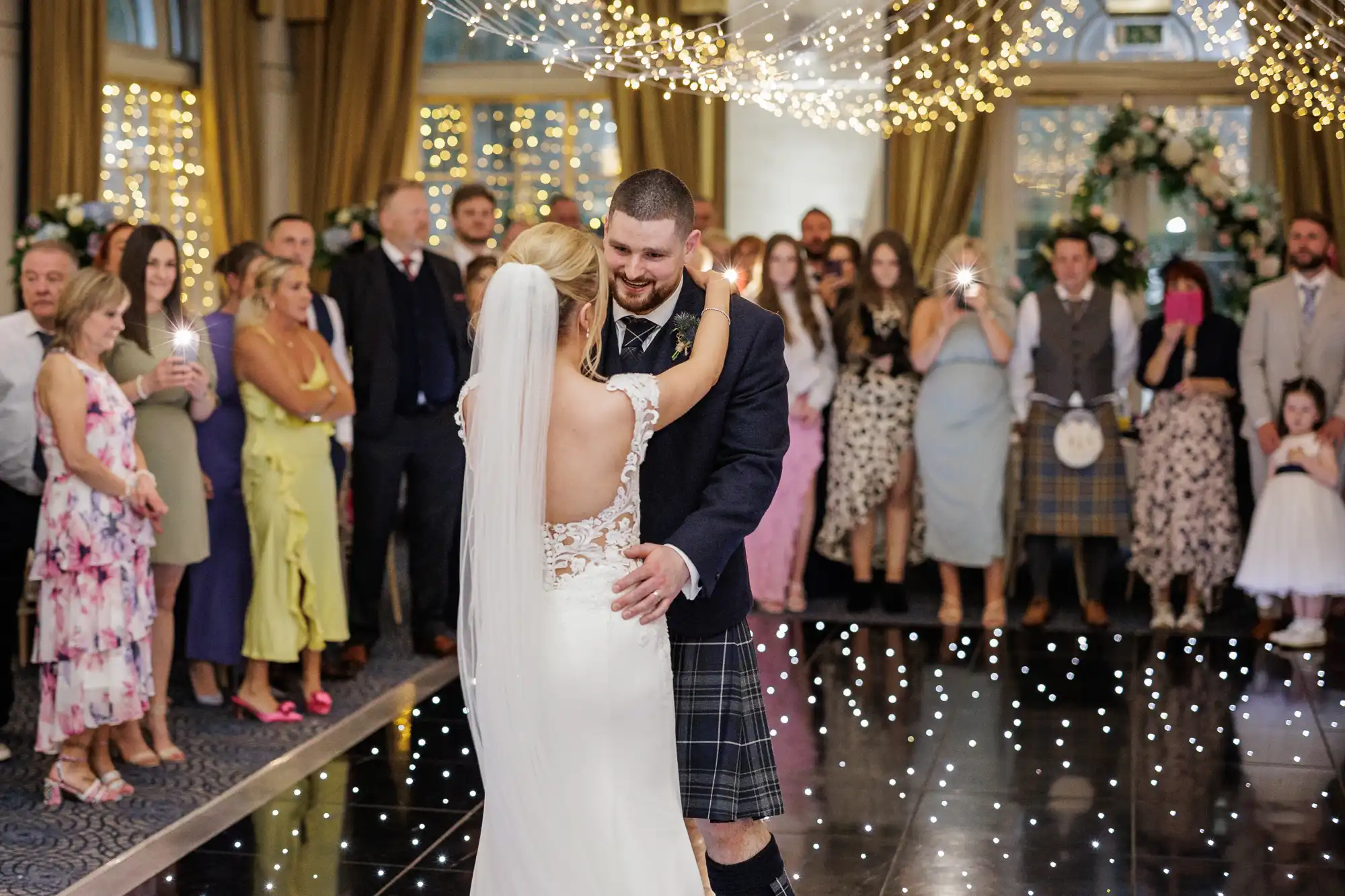 A bride and groom share their first dance under fairy lights, surrounded by guests in formal attire. The bride wears a white dress with lace details and a veil; the groom wears a kilt. Onlookers watch, some taking photos, in an elegant, decorated venue.