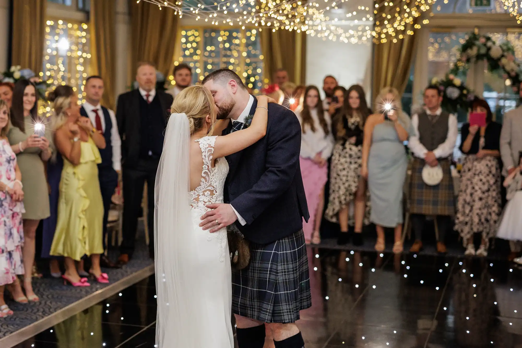 A bride in a white dress and veil and a groom in a jacket and kilt share a kiss on a glossy dance floor under fairy lights, surrounded by wedding guests dressed in formal and semi-formal attire, some taking photos.