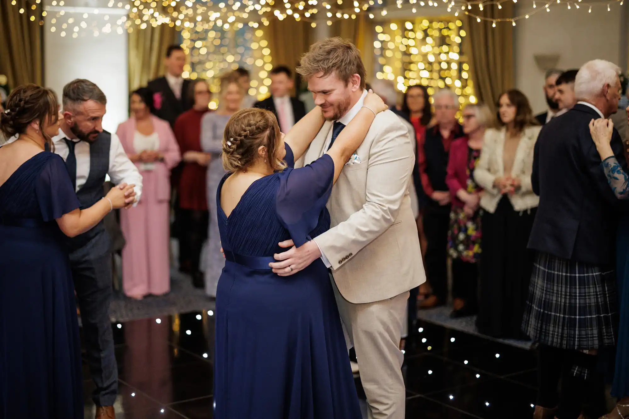 A couple dance together at a formal event, with the woman in a navy blue gown and the man in a beige suit. They smile at each other under strings of fairy lights. Other couples and guests, dressed formally, watch and dance in the softly lit background.