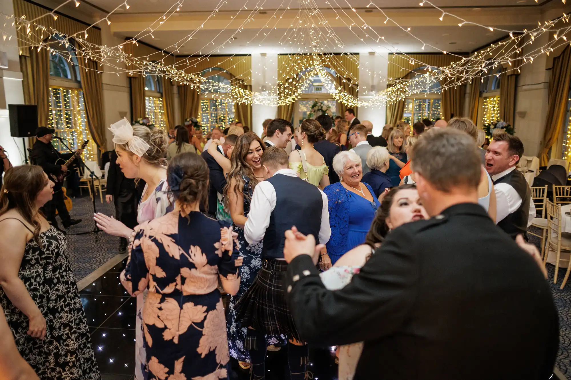 A lively wedding reception with guests dancing under strings of fairy lights in an elegant hall. People of various ages smile and interact, dressed in formal and semi-formal attire. The festive atmosphere is warm and joyful, with golden decorations.