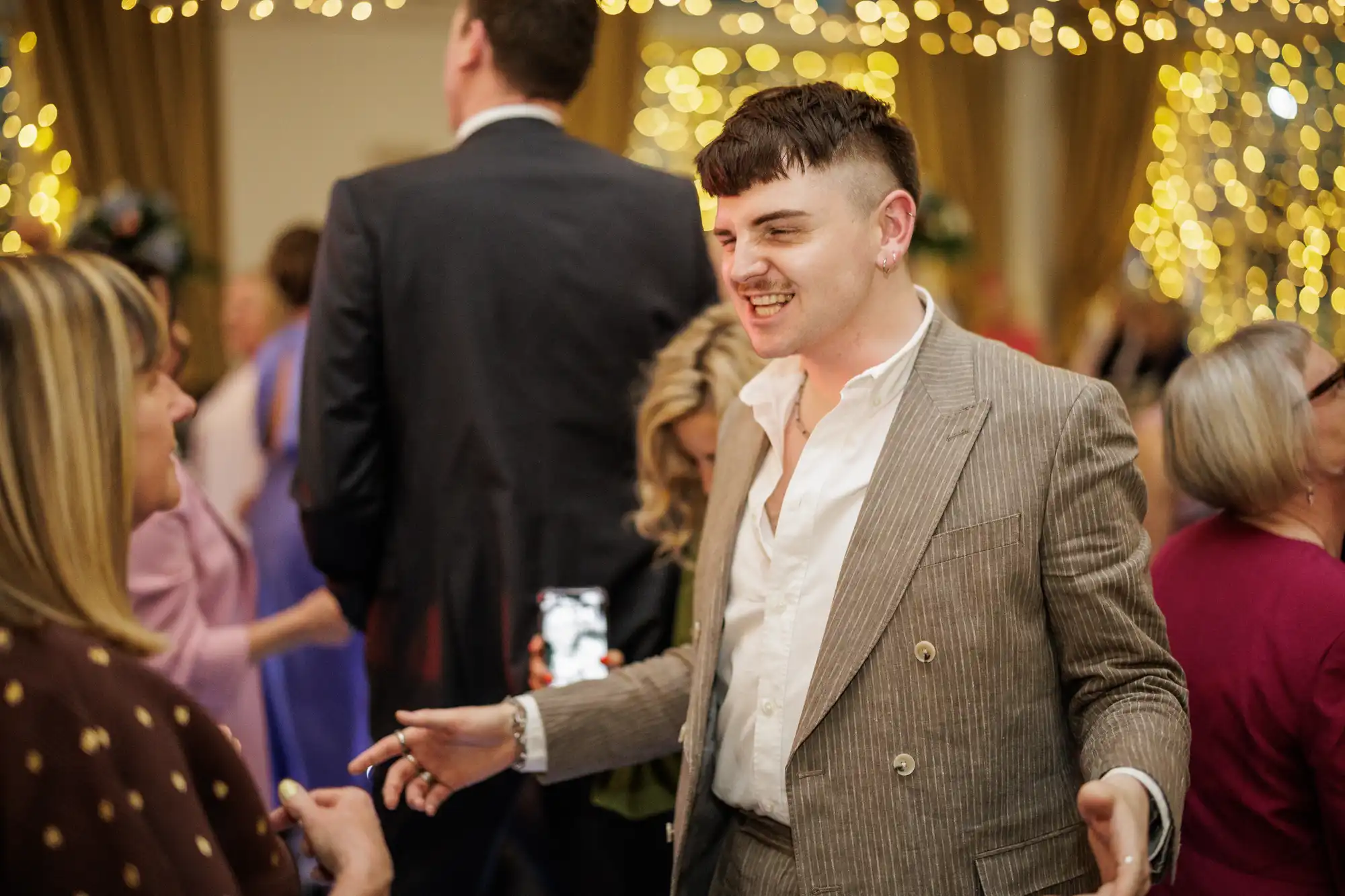 A young man in a pinstripe suit smiles and gestures whilst talking at a lively indoor event. Warm fairy lights hang in the background. Other people, dressed formally, are socialising around him. The atmosphere appears festive and cheerful.