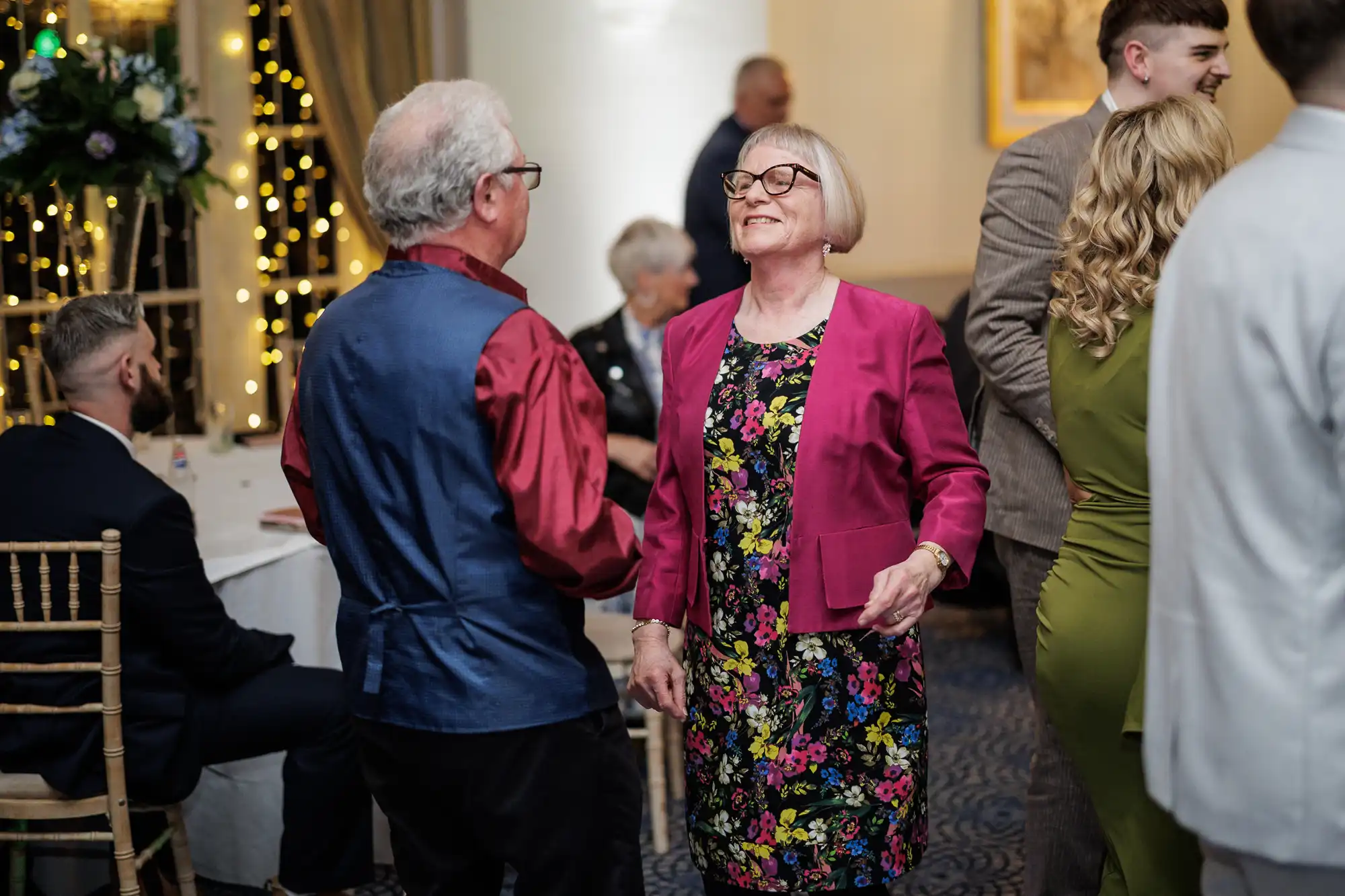 An older woman in glasses, a floral dress, and a pink blazer smiles whilst dancing with an older man in a red shirt and blue waistcoat at an indoor event. People sit and stand around, with fairy lights and formal decor in the background.