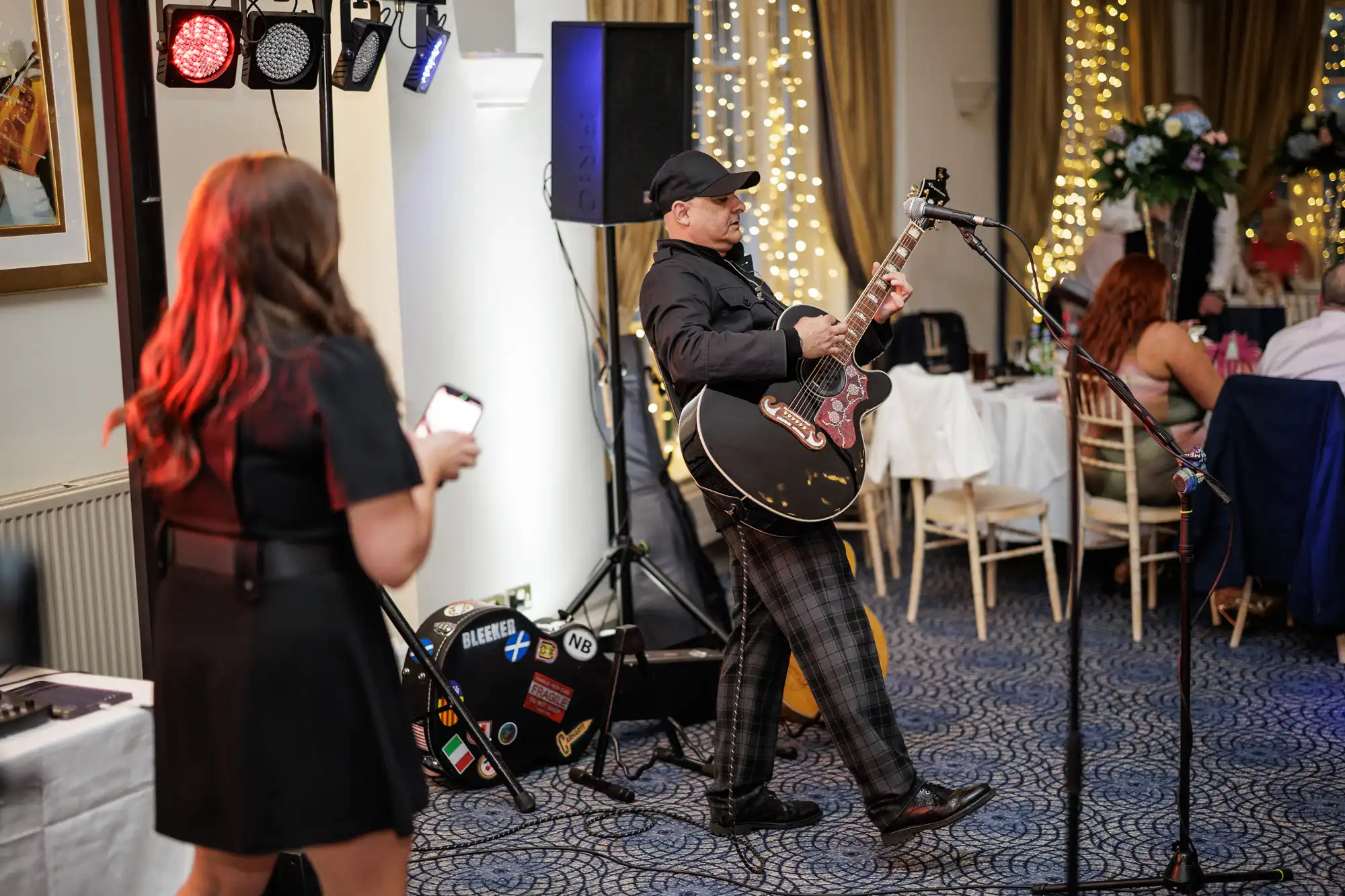 A musician wearing a black cap and checked trousers plays an acoustic guitar and sings into a microphone in a festive, decorated room. A woman in a black dress stands nearby holding a mobile. Warm fairy lights and seated guests are in the background.