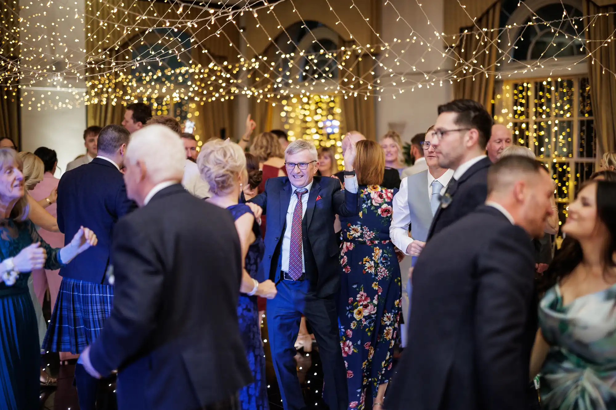 A lively group of people dressed formally dance and mingle at an indoor event under a ceiling decorated with strings of warm, glowing fairy lights. The atmosphere is festive, with many guests smiling and enjoying themselves on the dance floor.