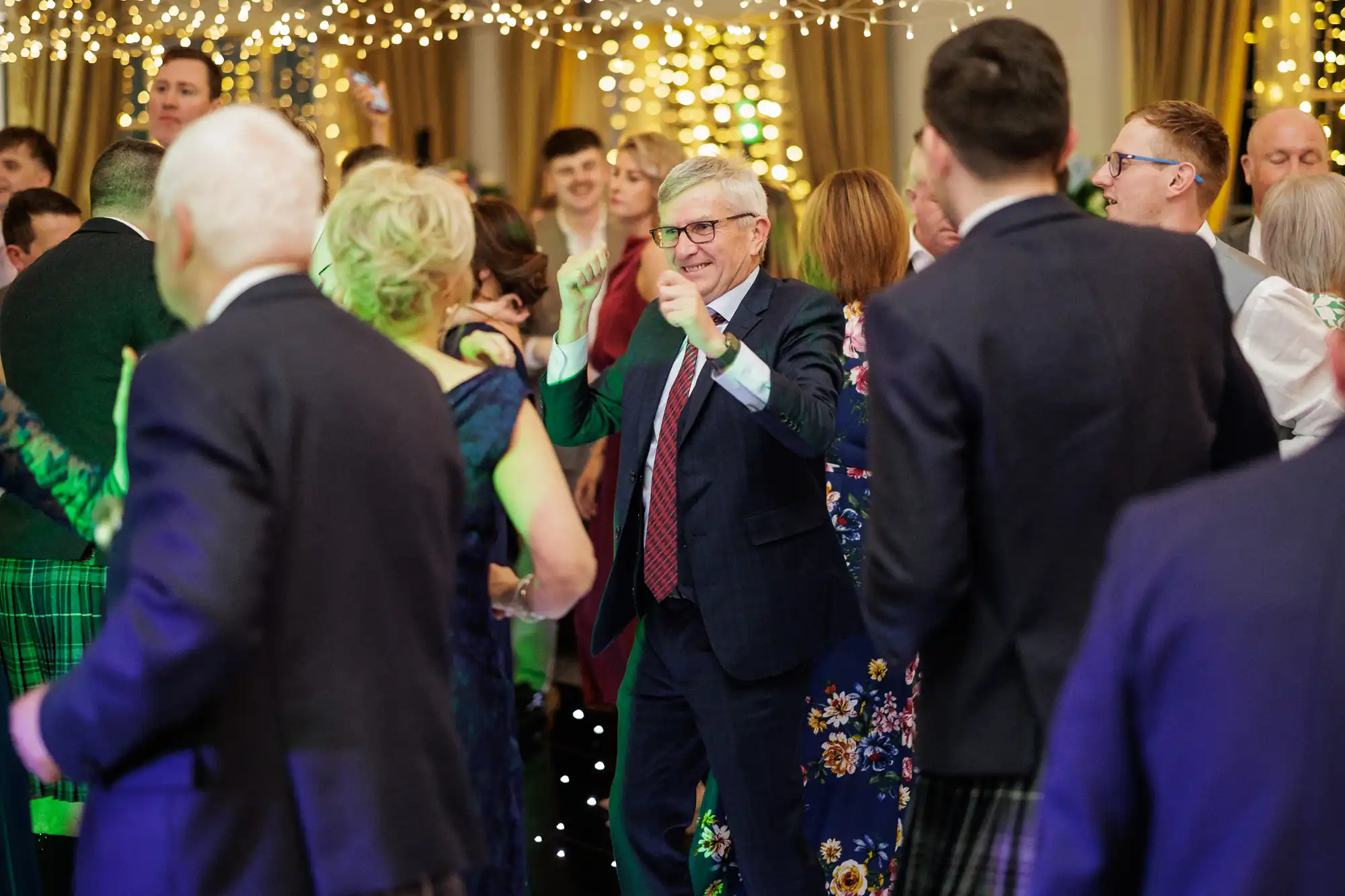 A group of people dressed in formal attire dance and enjoy themselves at a festive indoor event. Fairy lights hang above, creating a warm, celebratory atmosphere. In the centre, an older man in glasses and a suit smiles and raises his hands while dancing.