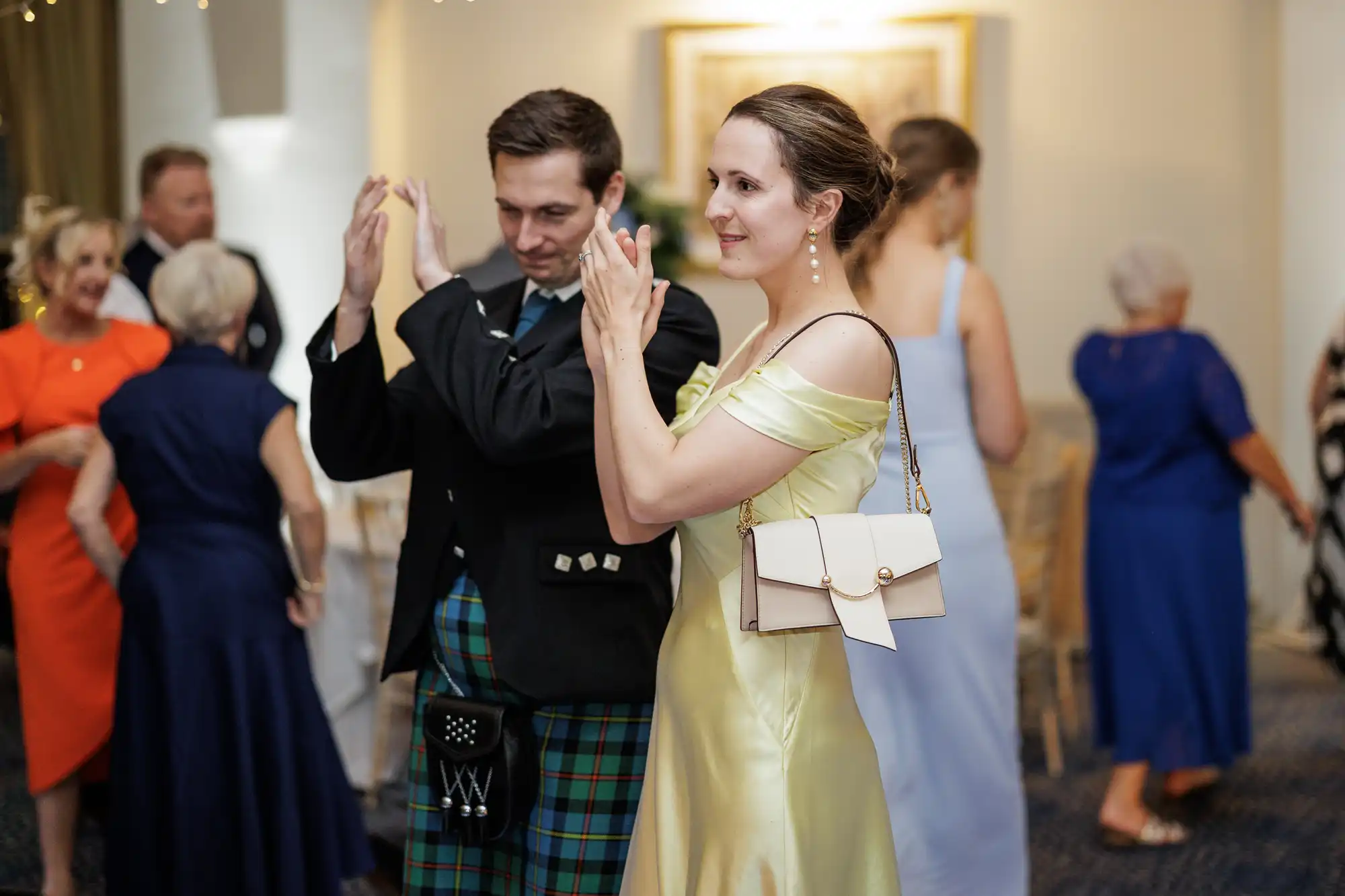 A man in a kilt and formal jacket and a woman in a yellow satin dress clap whilst dancing at an indoor event. Other guests in colourful dresses and suits mingle in the blurred background under warm lighting.