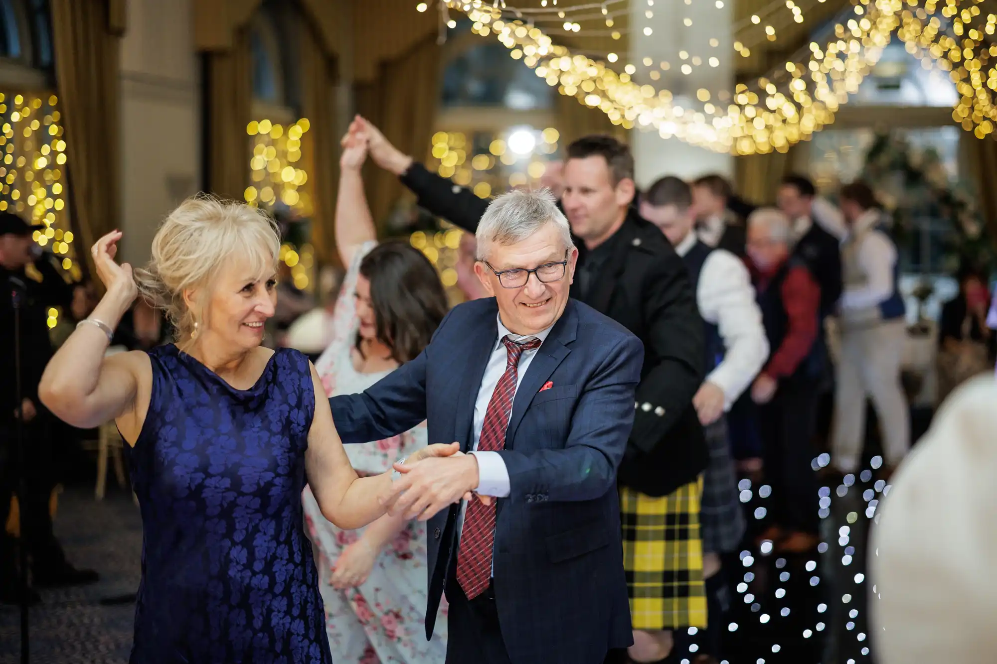 Older adults in formal attire dance together at a festive indoor event. Fairy lights hang overhead, creating a warm atmosphere. Other guests in the background, some in kilts, join the lively celebration under twinkling lights.