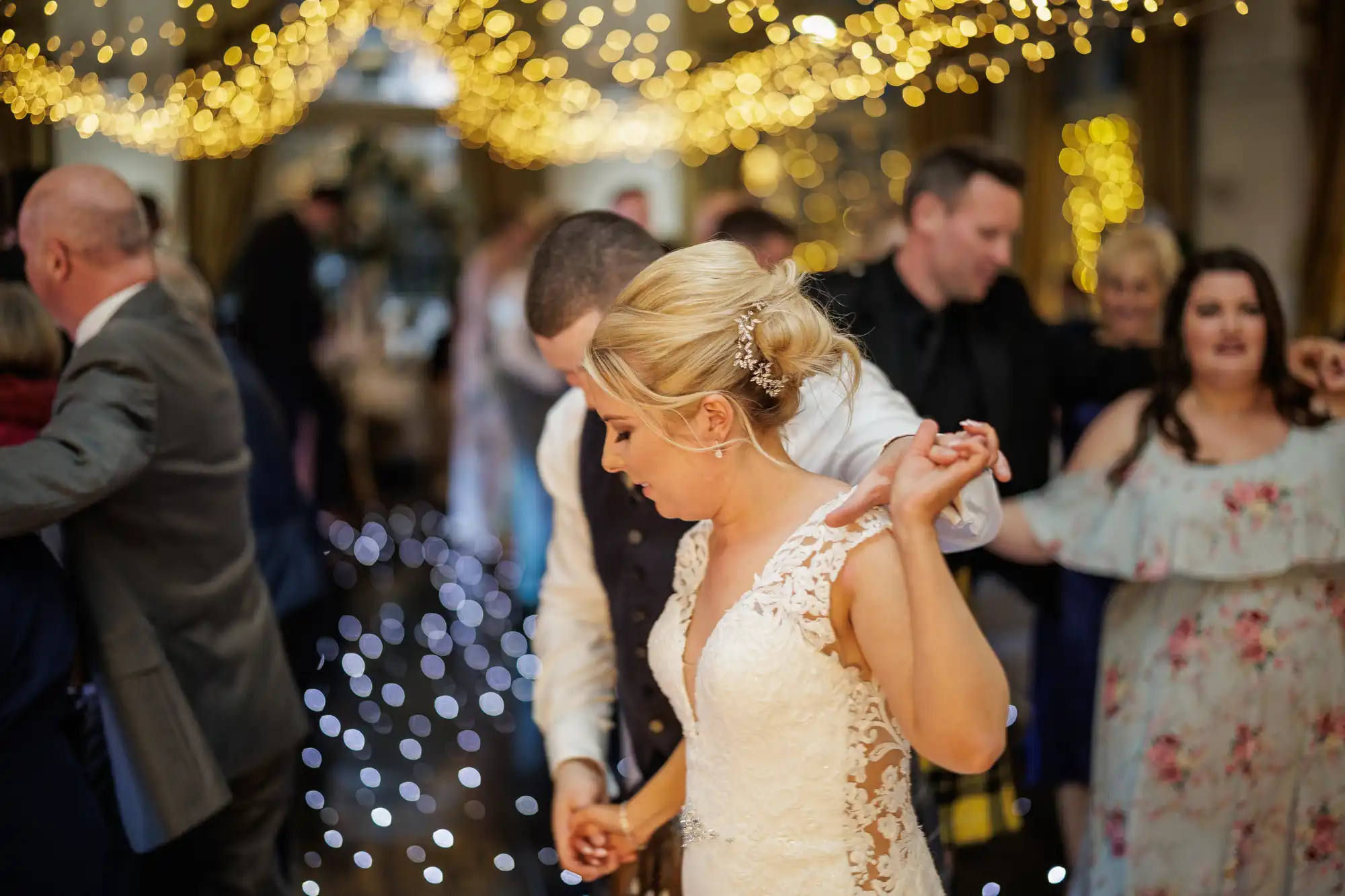 A bride in a lace wedding dress dances closely with a groom under warm, golden fairy lights. Guests in formal attire dance around them, creating a lively and joyful atmosphere at the wedding reception. The background is softly blurred with twinkling lights.