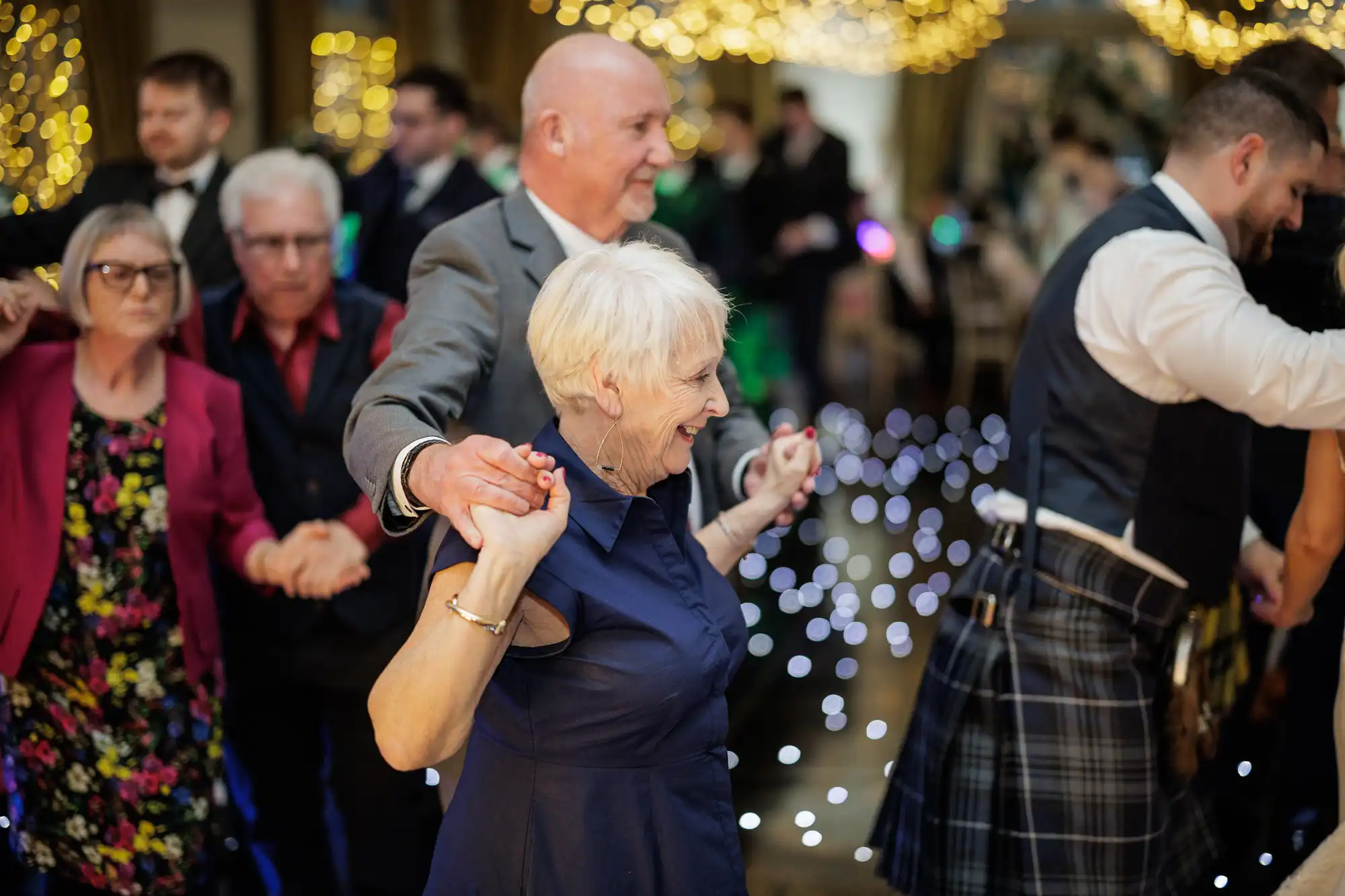 A group of people, mostly older adults, form a conga line and dance at a festive indoor event with fairy lights and bokeh in the background. The mood is joyful, and the setting appears to be a lively celebration or party.