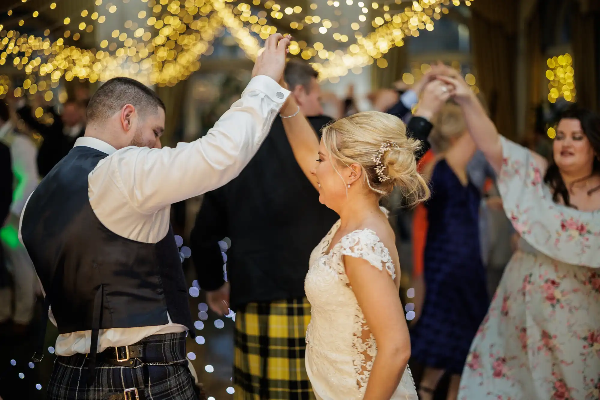 A bride in a white lace gown and a groom in a kilt and waistcoat dance together under glowing fairy lights, holding hands and smiling. Other guests in festive attire dance in the background, creating a lively wedding celebration atmosphere.