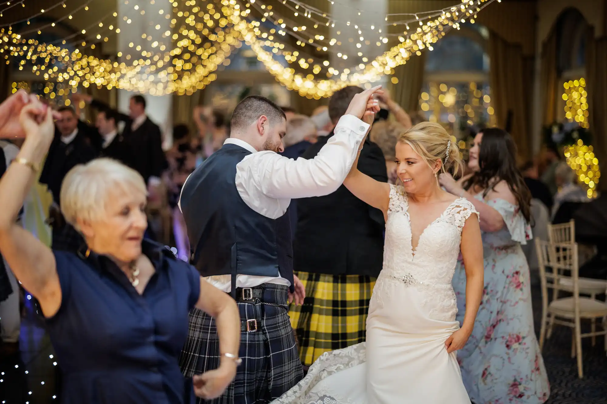 A bride in a white lace dress and a groom in a waistcoat and kilt dance under strings of warm fairy lights, smiling. Guests, including an older woman in blue and people in tartan kilts, dance around them in a festive, warmly lit banqueting hall.