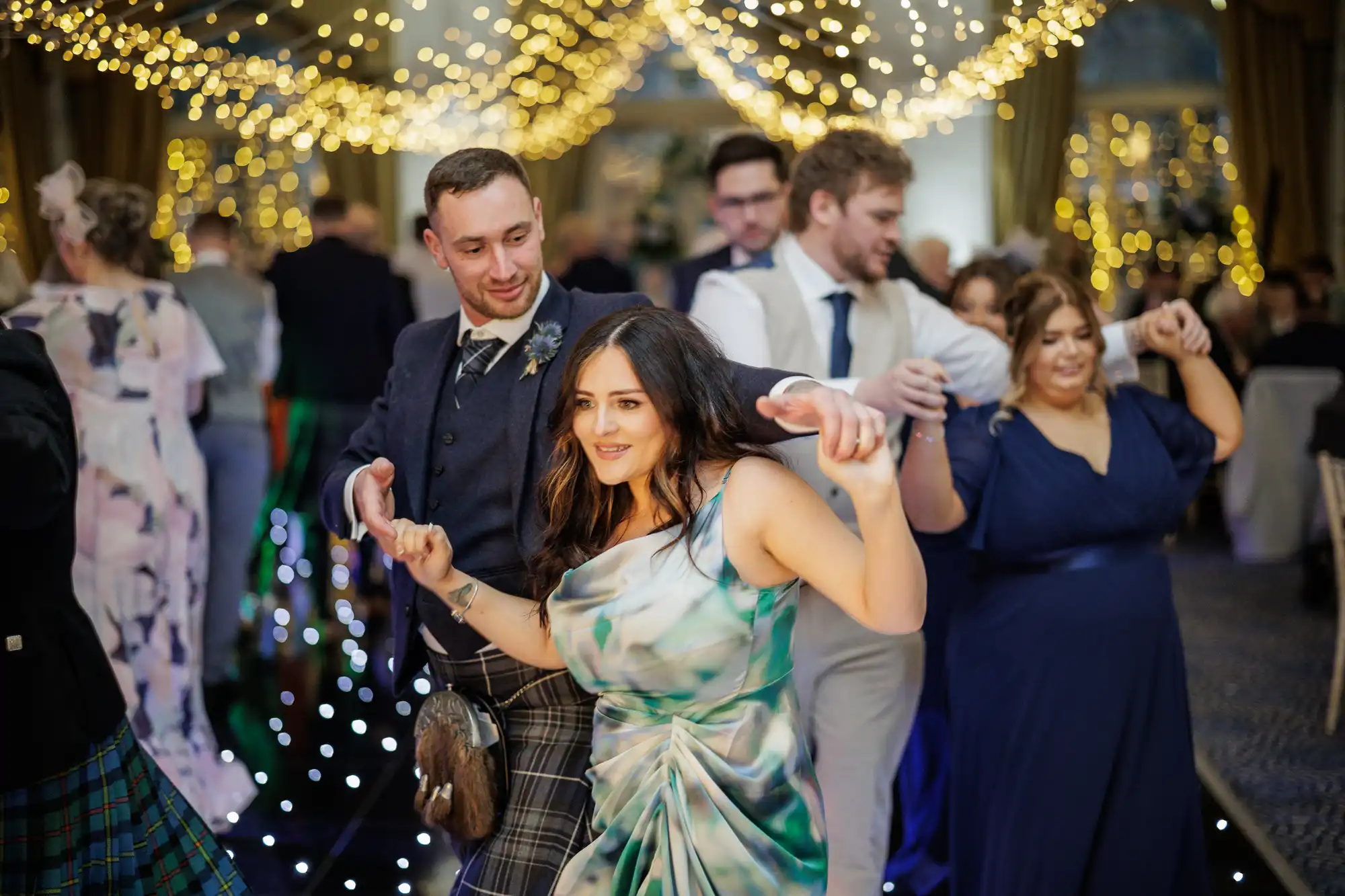 A group of people dance joyfully under fairy lights at a formal event. A woman in a green patterned dress leads, smiling, with a man in a suit behind her. Other guests in formal attire are dancing in the warmly lit, decorated venue.