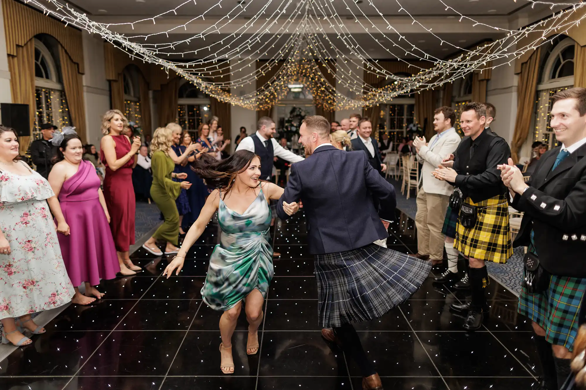 A lively group dances on a shiny black floor under strings of fairy lights. A woman in a green dress and a man in a blazer and kilt joyfully lead, surrounded by clapping guests in colourful dresses and kilts, in an elegant hall with tall windows.
