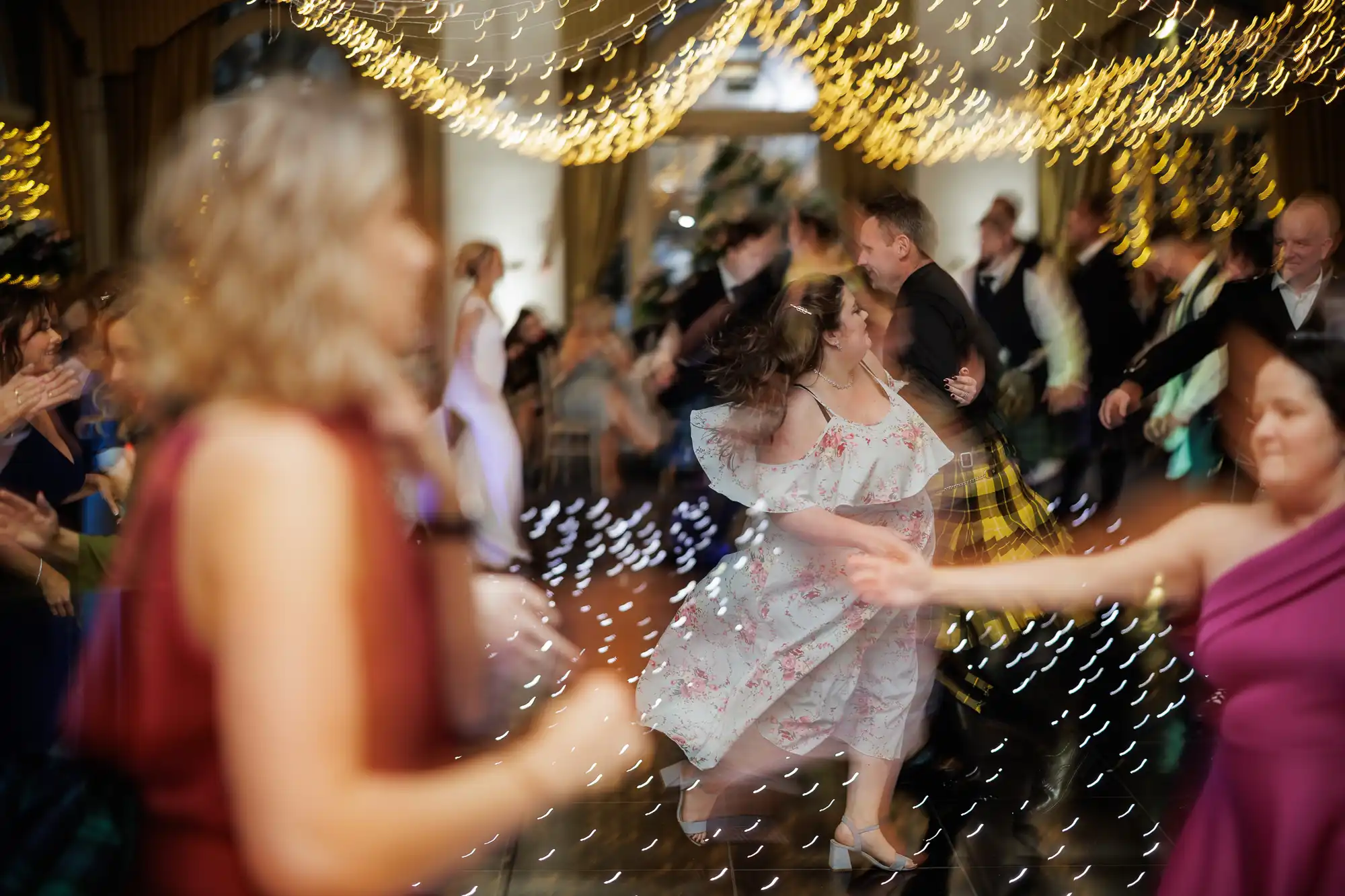 A lively dance scene with couples spinning under warm fairy lights. Motion blur captures the movement and energy. People wear formal or semi-formal attire, with one woman in a floral dress twirling at the centre. The background is a warmly lit, festive room.
