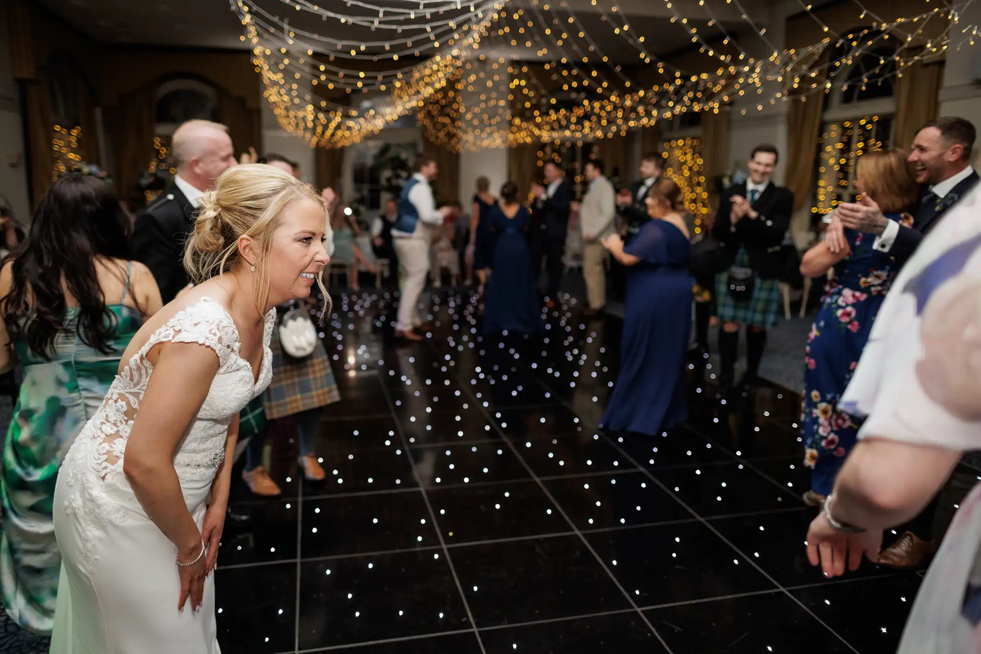 A bride in a white dress leans forward, smiling and laughing on a black, star-lit dance floor. Guests in formal attire dance and mingle under twinkling fairy lights, creating a festive, joyful atmosphere at an indoor wedding reception.