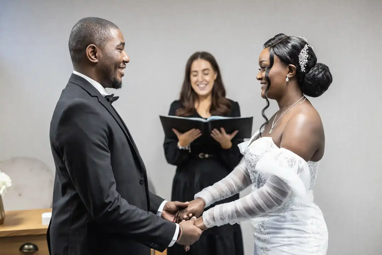 A bride and groom stand facing each other, smiling and holding hands. The bride wears a white dress and gloves, the groom a black tuxedo. A woman in black stands behind them, holding a book and smiling, likely officiating the wedding ceremony.
