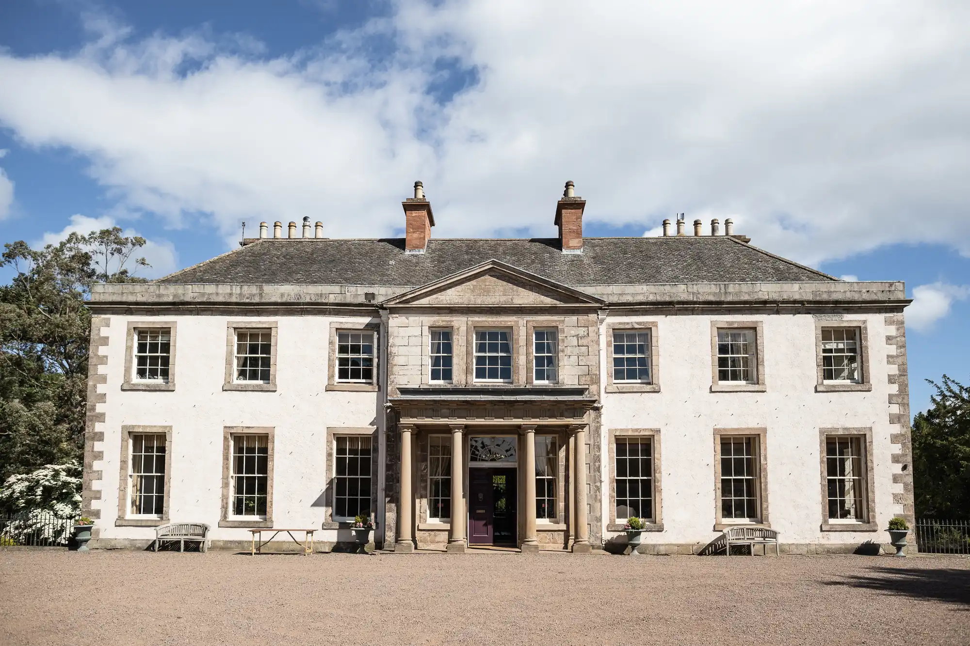 A large, symmetrical Georgian-style manor house with pale stone walls, rows of tall sash windows, and a central entrance framed by columns. The building is set against a blue sky with clouds and surrounded by gravel and greenery.
