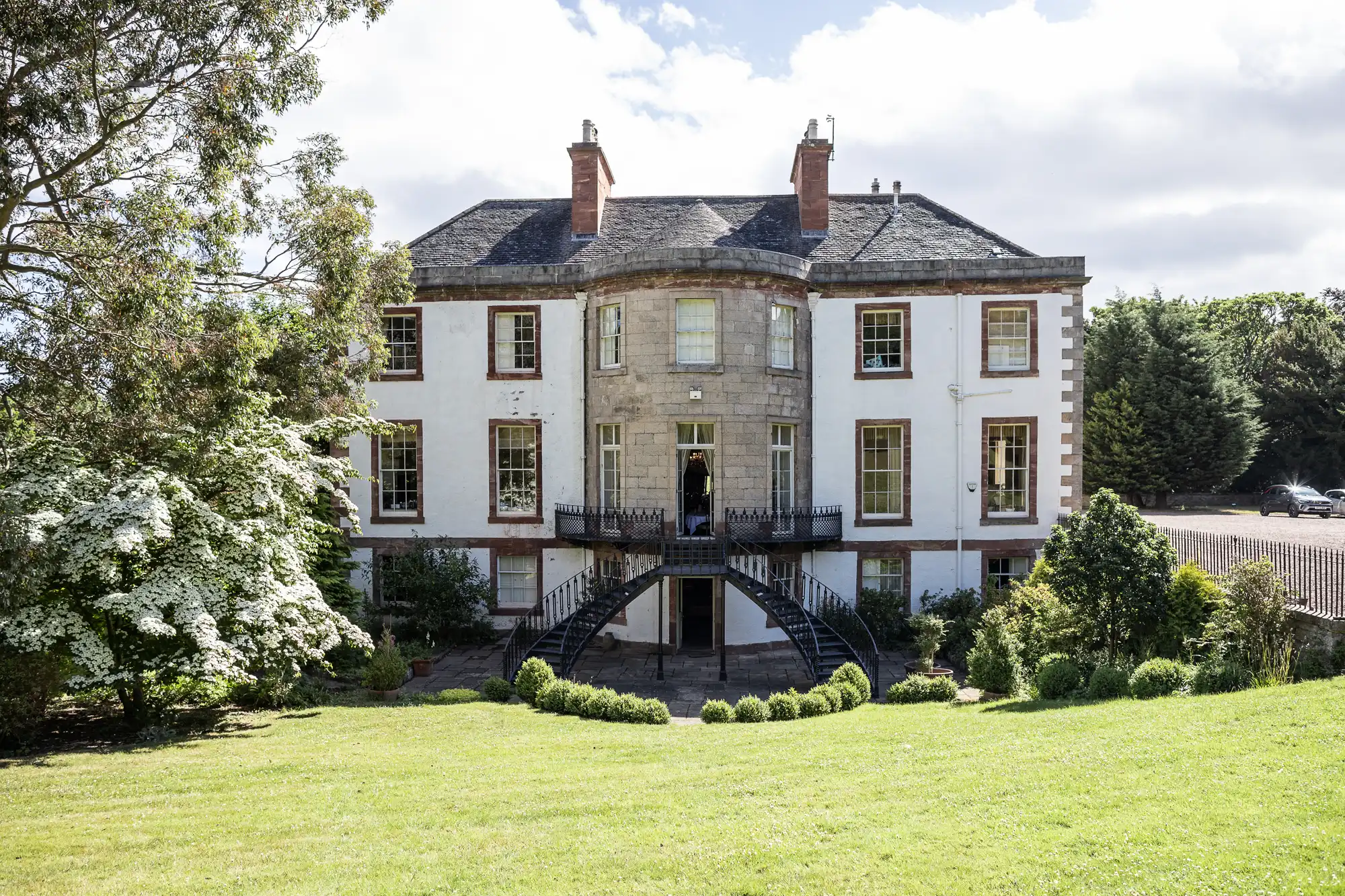 A large, elegant Georgian-style mansion with white walls, tall sash windows, and a central rounded bay with double staircases leading to a grassy lawn, surrounded by lush trees and landscaped gardens under a partly cloudy sky.
