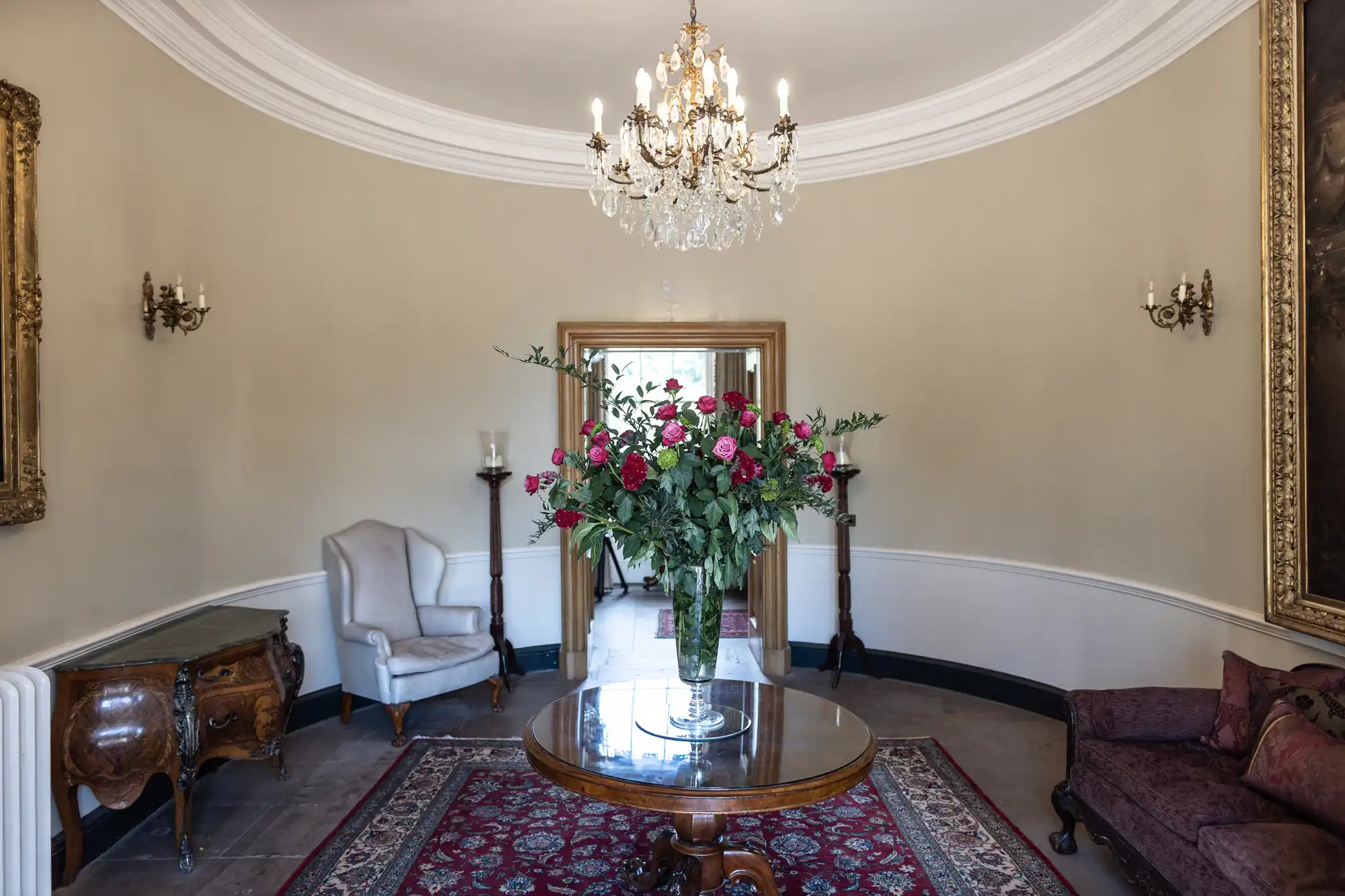 Elegant, round room with a chandelier, beige walls, and a large ornate mirror. A glass vase of pink and red flowers sits on a round wooden table atop a patterned rug. Antique furniture, including armchairs and a sofa, lines the walls.
