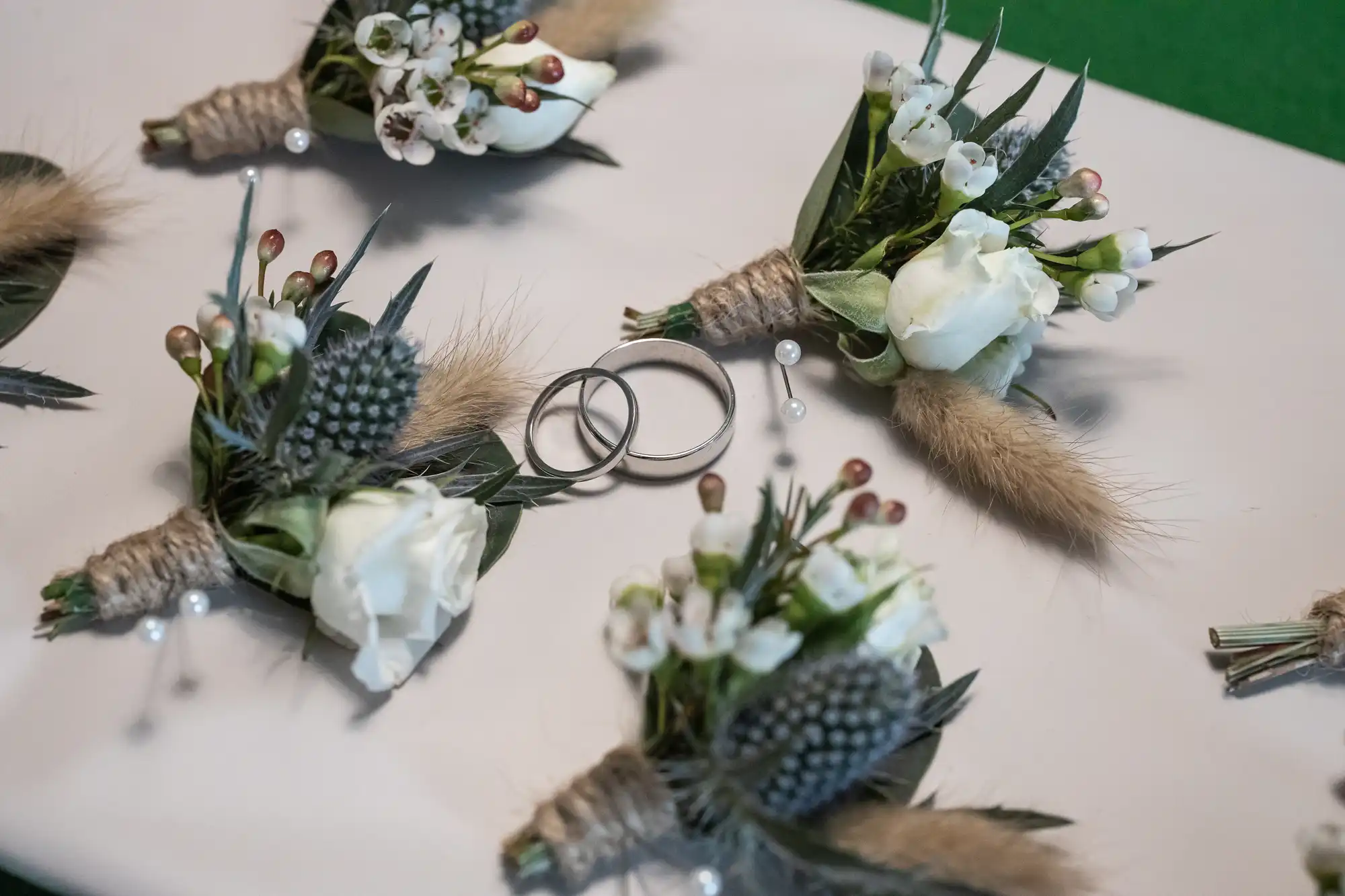 Two silver wedding rings rest on a white surface, surrounded by small white floral buttonholes with greenery, thistle, and wrapped twine stems. Decorative pins with white pearl heads accompany the arrangements.