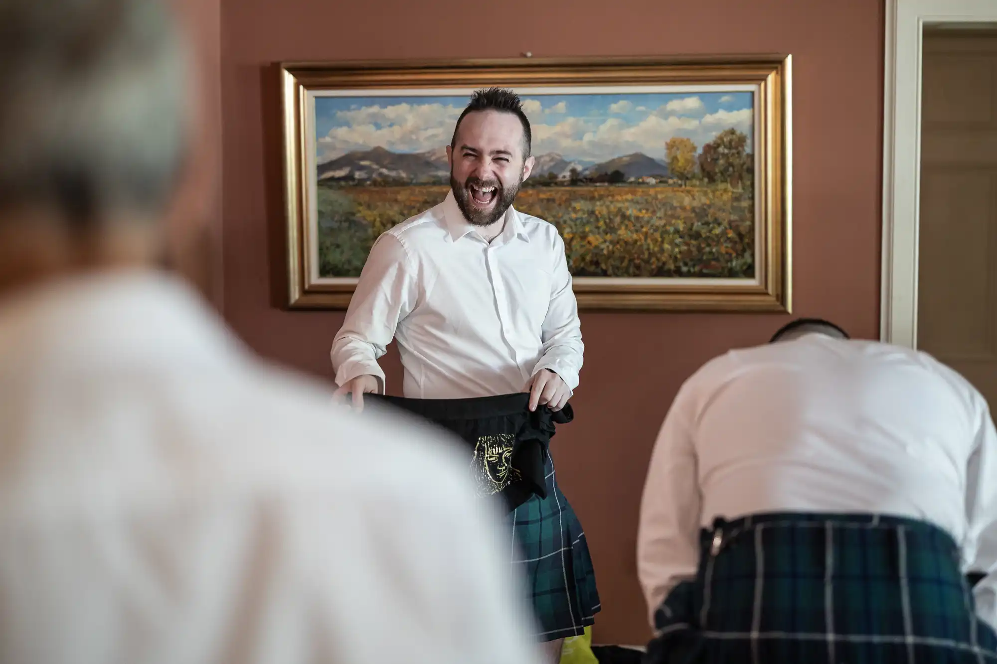 A man smiling widely holds a kilt in a warmly lit room with brown walls. Two other people, partially visible and also wearing kilts, stand nearby. A landscape painting with mountains and trees hangs on the wall behind them.