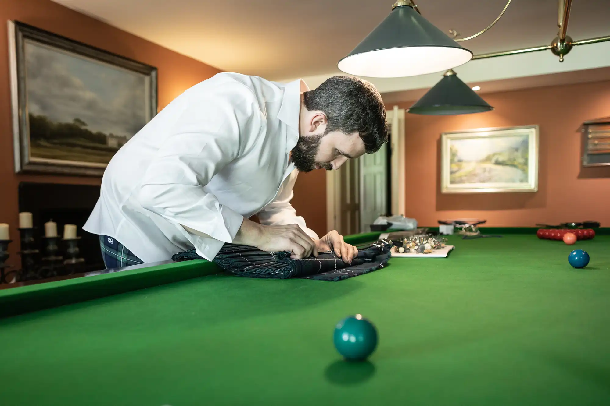 A bearded man wearing a white shirt and blue jeans leans over a green snooker table, carefully working on a folded black cloth. Several snooker balls, a cue, and a lamp above the table are visible, with paintings and furniture in the background.