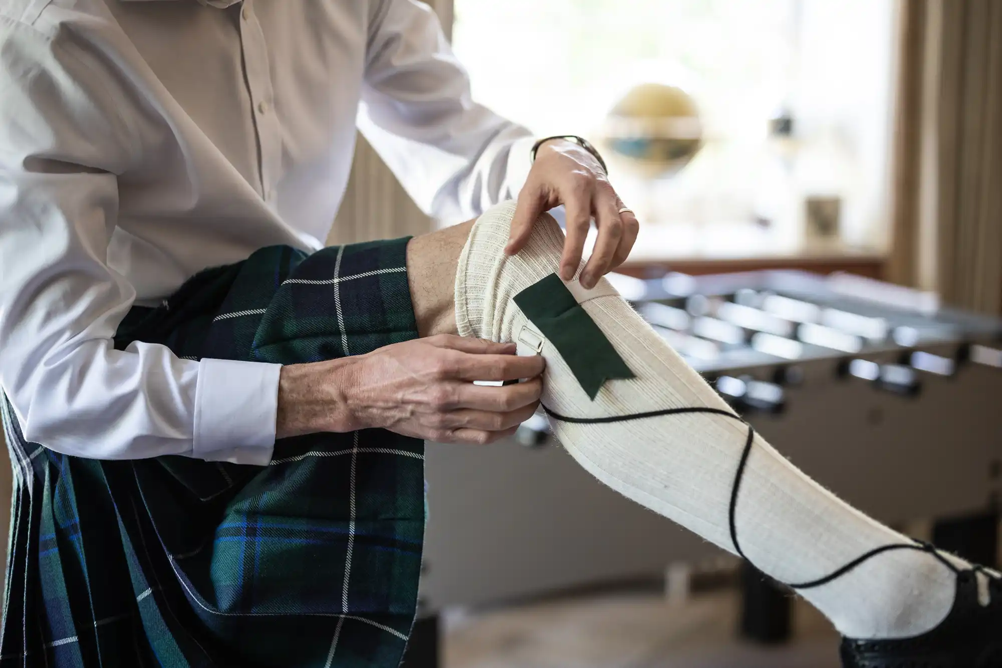 A person in a white shirt and tartan kilt adjusts a cream-coloured knee-high sock with a green garter or ribbon. The individual sits next to a table football table in a well-lit room, focusing on preparing traditional Scottish attire.