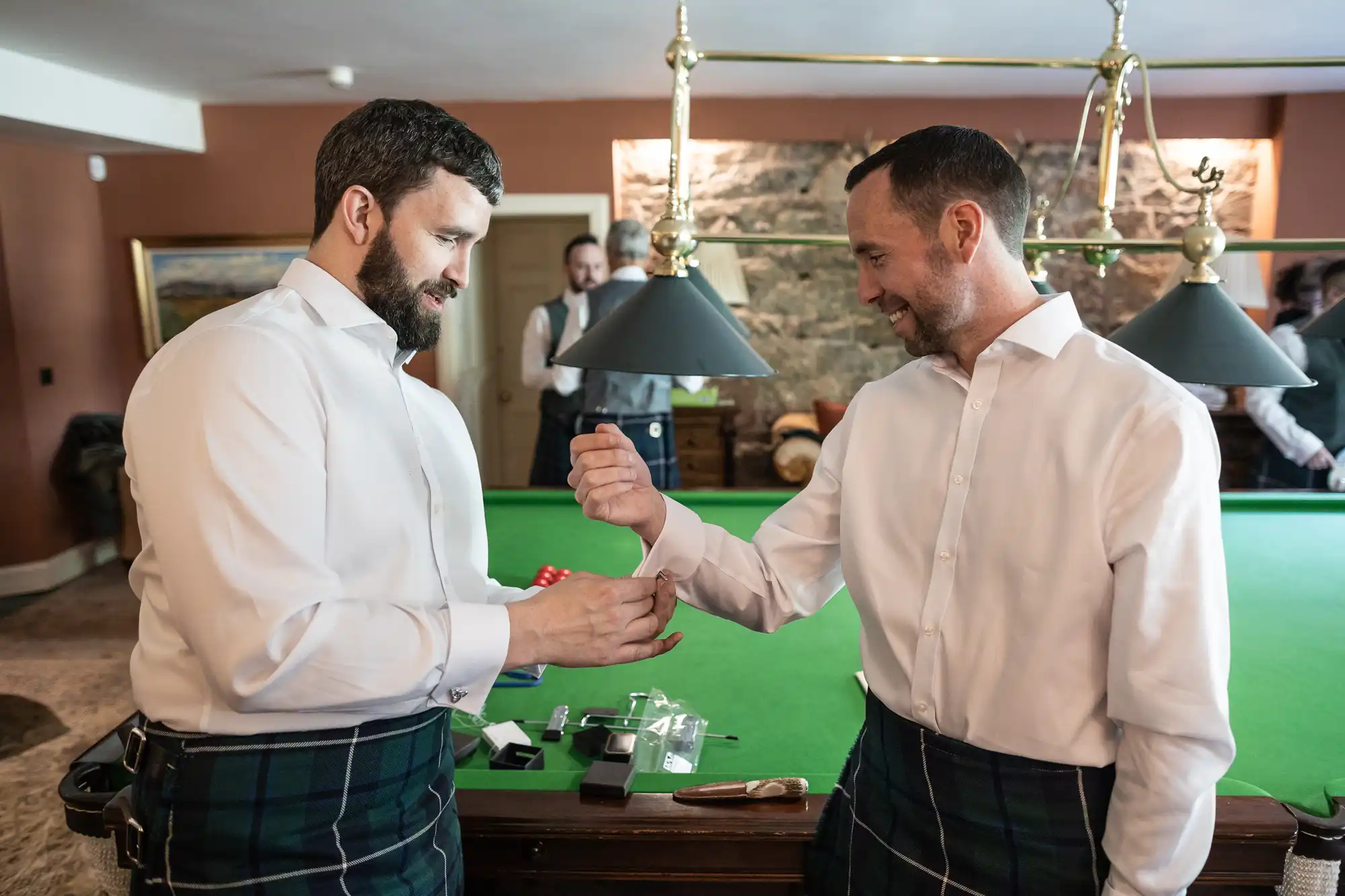 Two men in white shirts and tartan kilts stand by a pool table, smiling as one fastens the other's cufflink. Others in similar attire chat in the background. The room has a stone wall, overhead lights, and scattered grooming items on the green table.