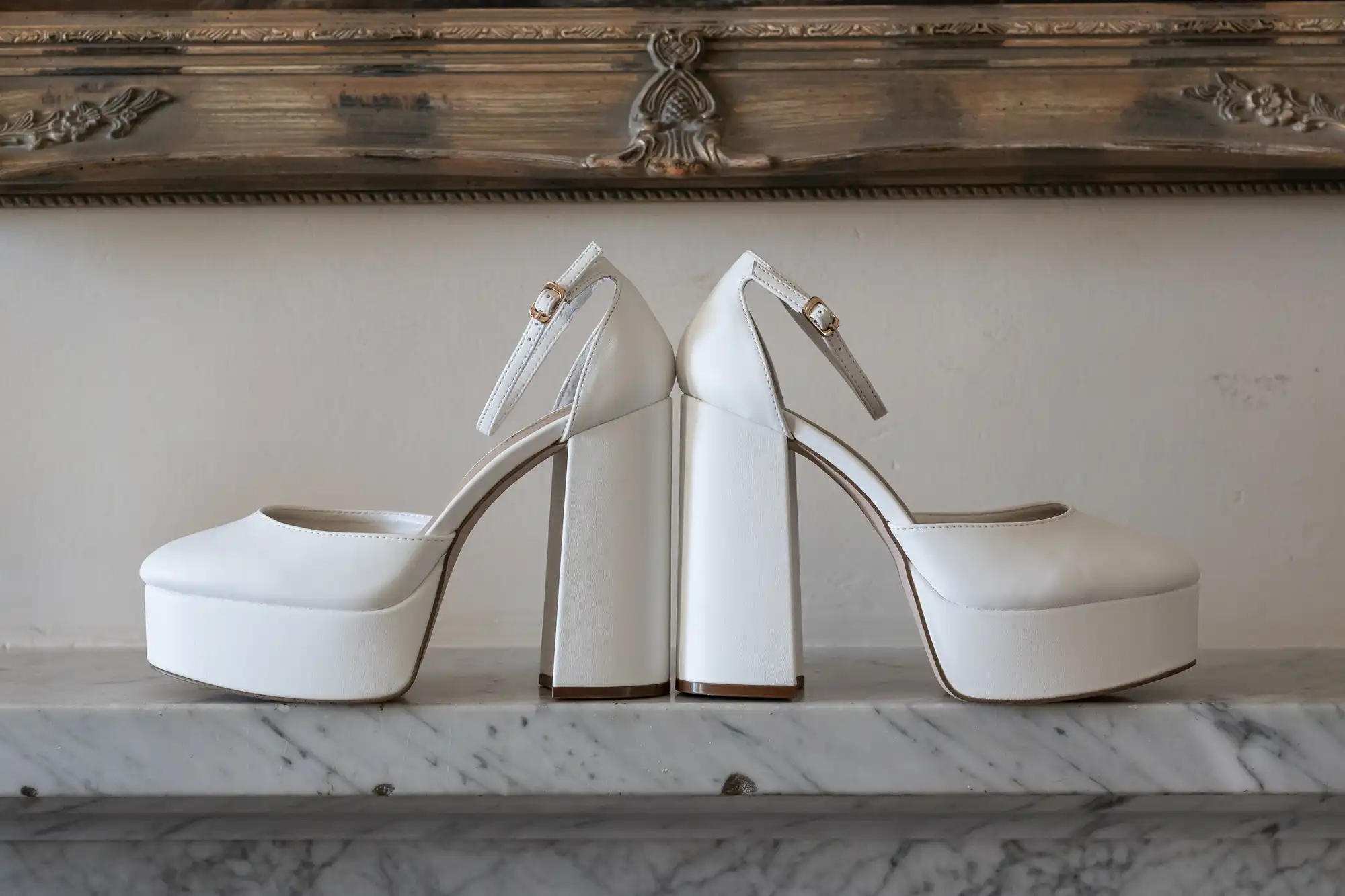 A pair of white high-heeled platform shoes with ankle straps sits side by side on a marble surface, in front of an ornate wooden frame or mantelpiece against a plain white wall.