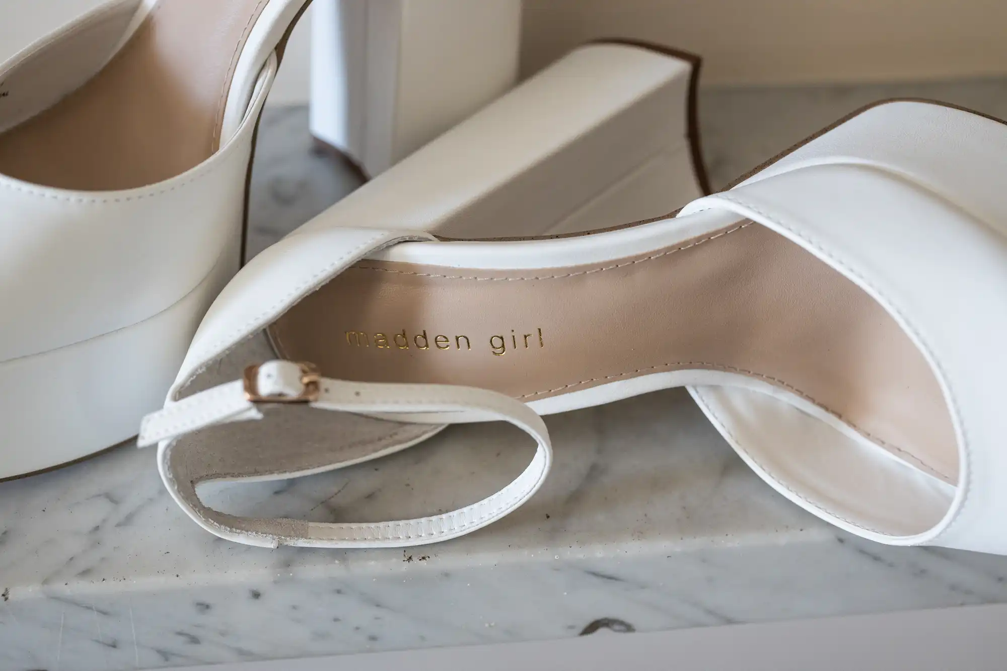 Close-up of a pair of white high-heeled platform sandals on a marble surface, with "madden girl" printed in gold lettering on the insole. One sandal is lying on its side, showing the buckle ankle strap and cushioned footbed.