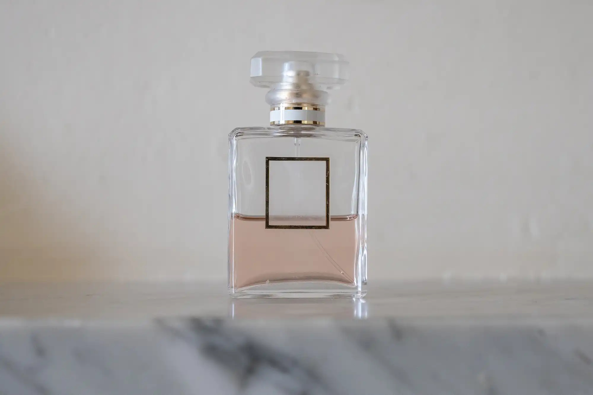 A clear glass perfume bottle with a square gold label and light pink liquid sits on a marble surface against a plain white background. The bottle has a faceted, translucent cap.