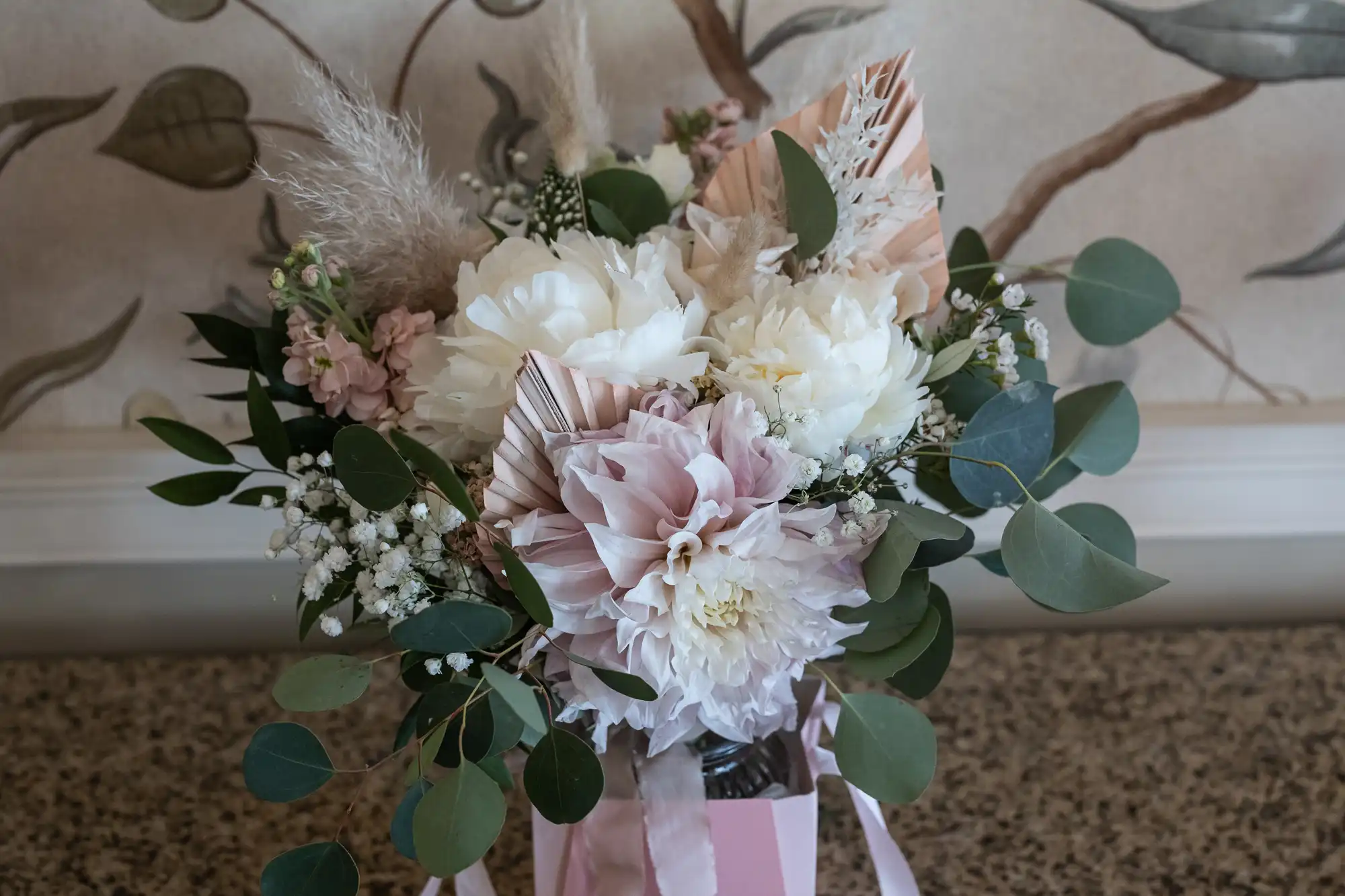 A floral arrangement features white and pale pink flowers, including dahlias and peonies, with greenery and dried leaves. The bouquet is displayed in a pink box on a speckled worktop, against a wallpapered background with leafy designs.
