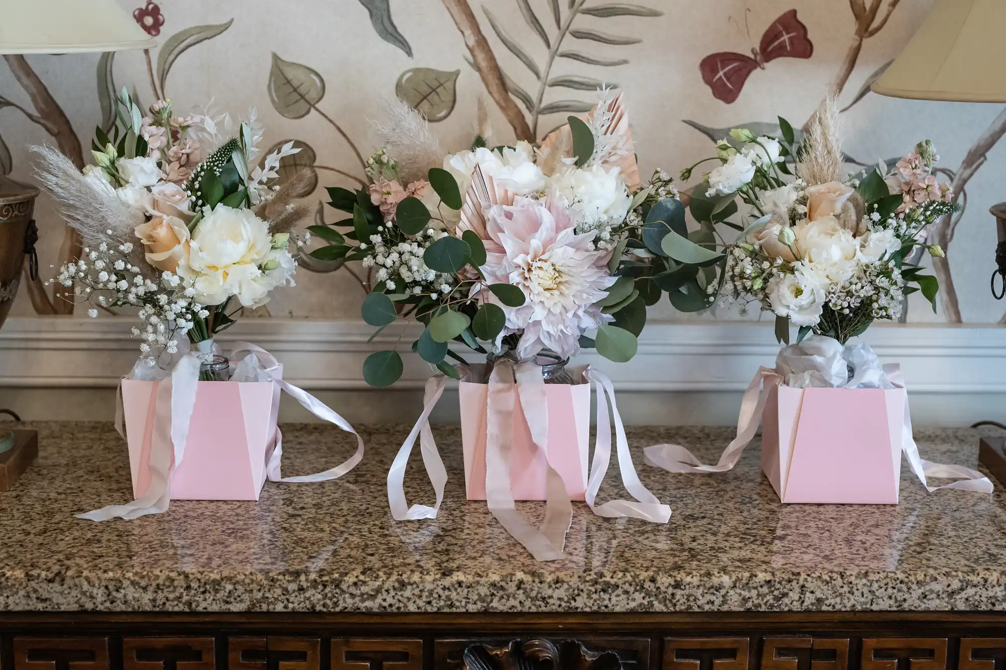 Three flower arrangements in pink boxes with white and blush flowers, greenery, and long pastel ribbons sit on a granite counter. Behind them is a wall with botanical wallpaper and two lamps partially visible on either side.