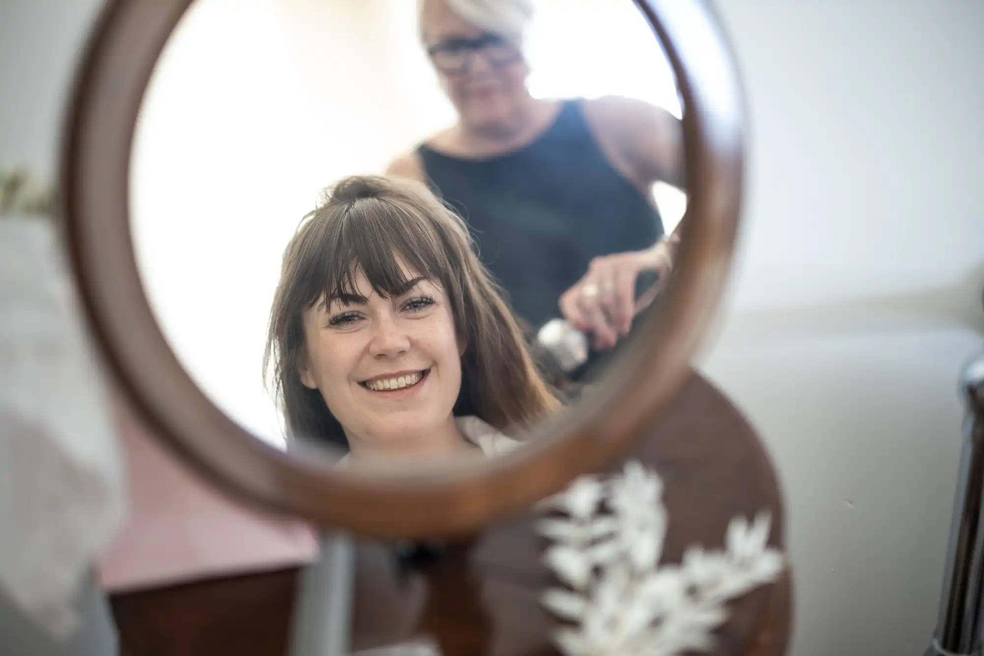 A woman with brown hair and a fringe smiles into a round mirror, her reflection showing her face clearly. Behind her, a person with short, fair hair and glasses styles her hair. The scene is indoors with soft lighting and blurred background objects.