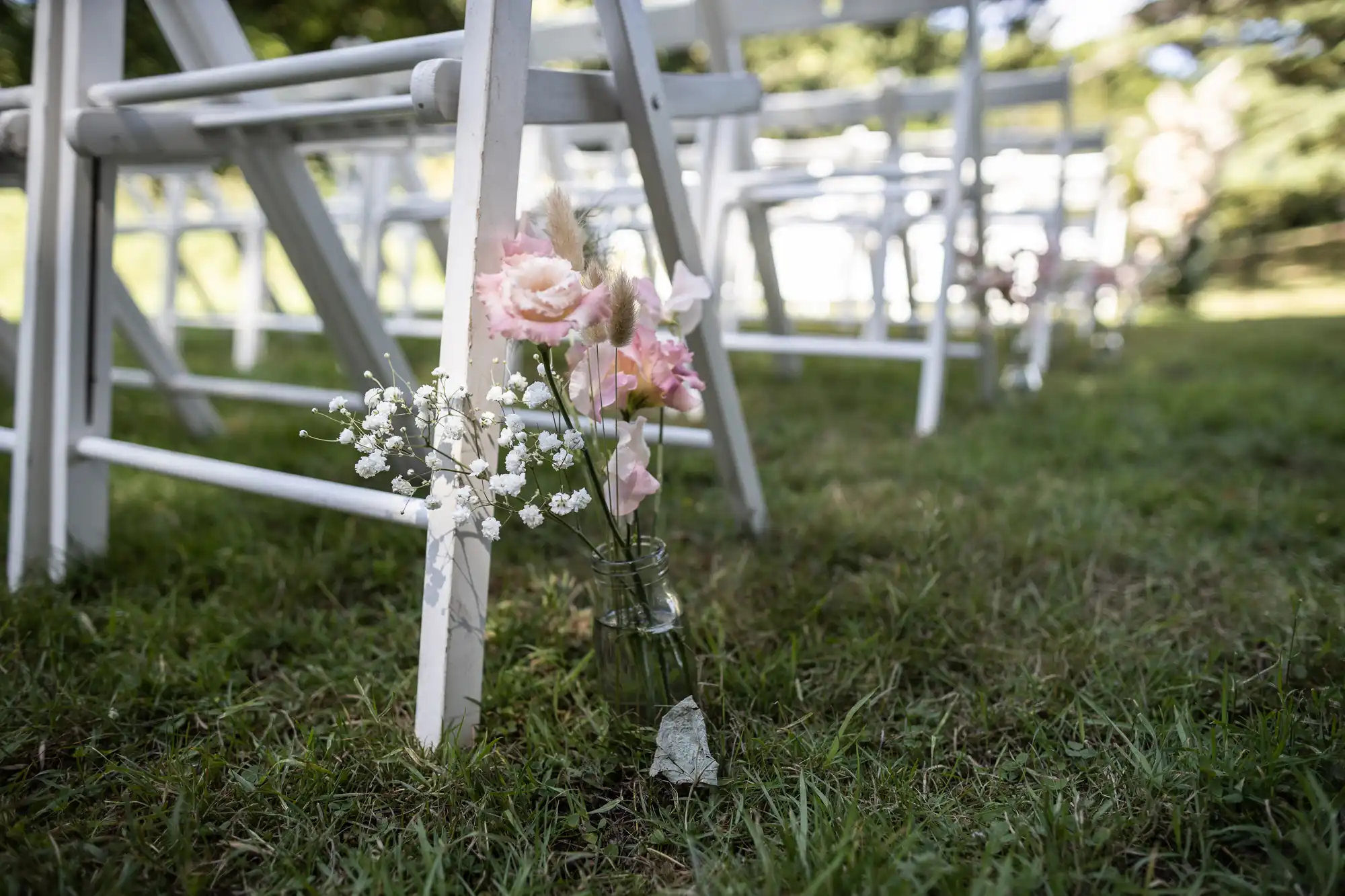 A small glass jar with pink roses and white baby's breath flowers is tied to a white wooden chair on green grass. The chair is part of a row set up outdoors, likely for a wedding or ceremony, with sunlight filtering through trees in the background.