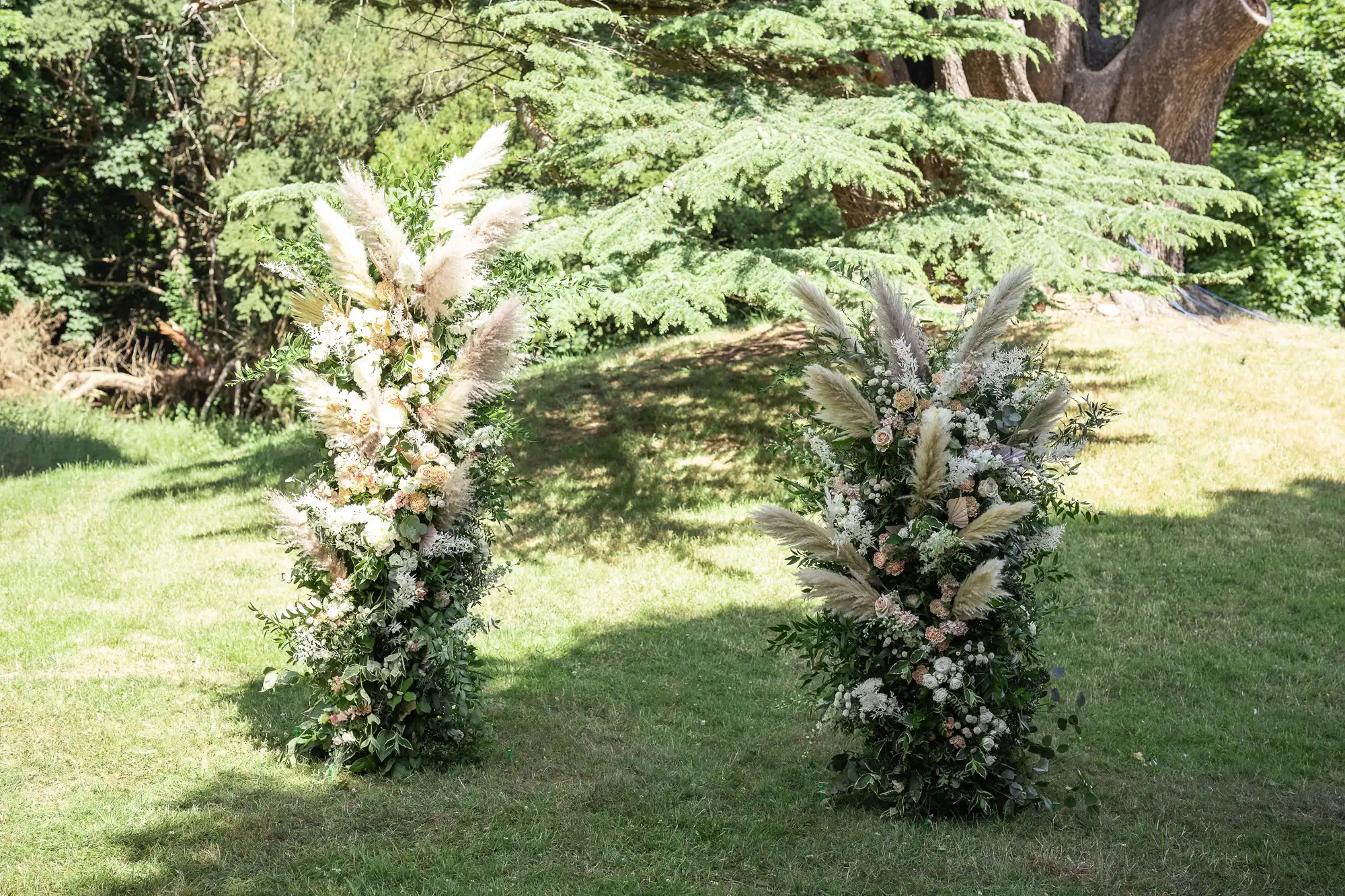 Two tall floral arrangements on a grassy lawn feature pampas grass, white and blush flowers, and greenery. Sunlight filters through trees in the background, casting dappled light and shadows across the lush outdoor setting.