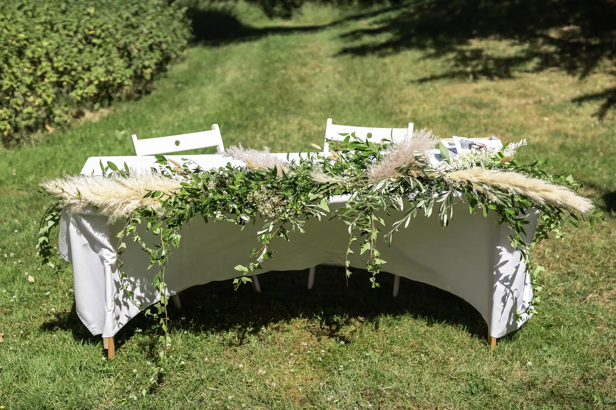 A white table covered with a white cloth is decorated with a lush garland of greenery and pampas grass. Two white chairs sit behind it on green grass, with trees and bushes in the background under bright sunlight.
