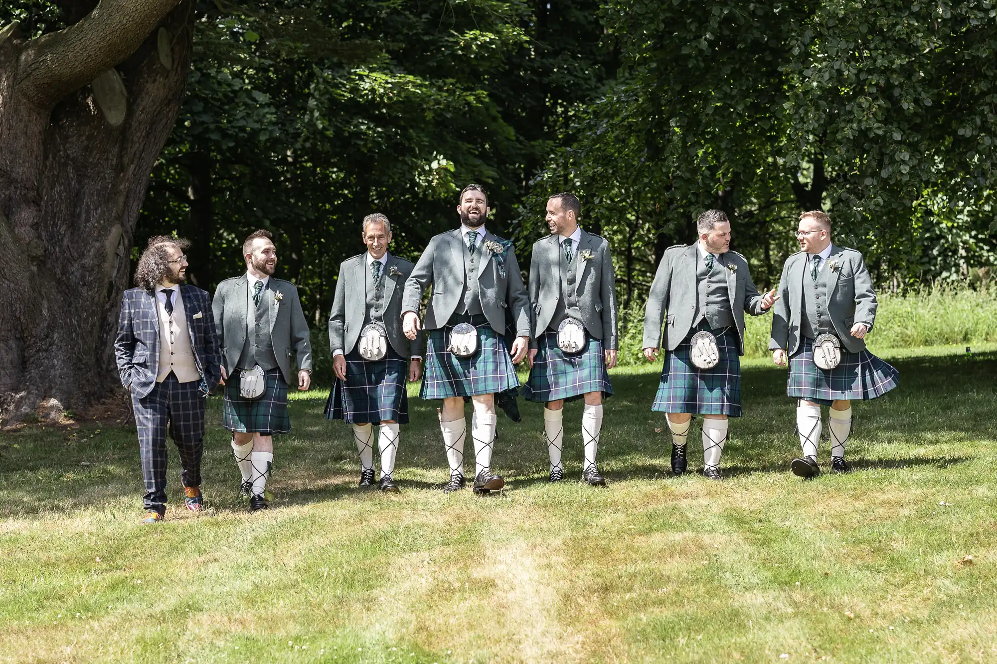 Seven men walk on grass in front of tall green trees. Six wear matching green jackets, tartan kilts, sporrans, and knee-high socks, while one on the left wears a checked suit with a waistcoat and tie. They all smile and appear to be enjoying themselves outdoors.
